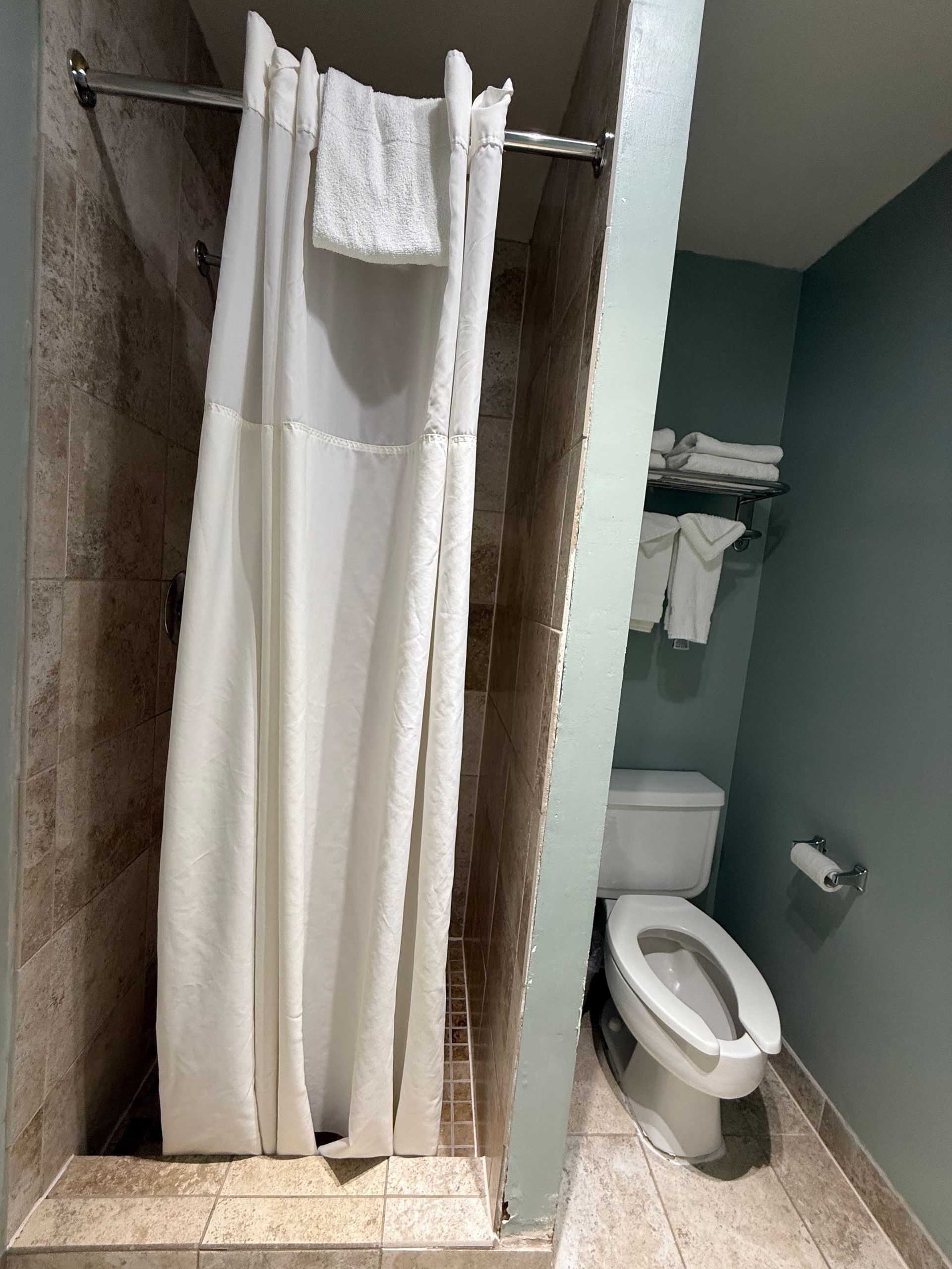 Shower with white curtain and towel, next to a toilet with a towel rack in a small bathroom with light green walls.