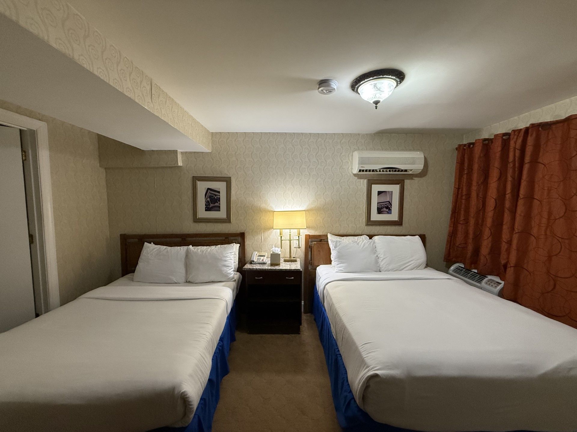 Two beds in a hotel room with nightstand, lamps, pictures, and curtains.