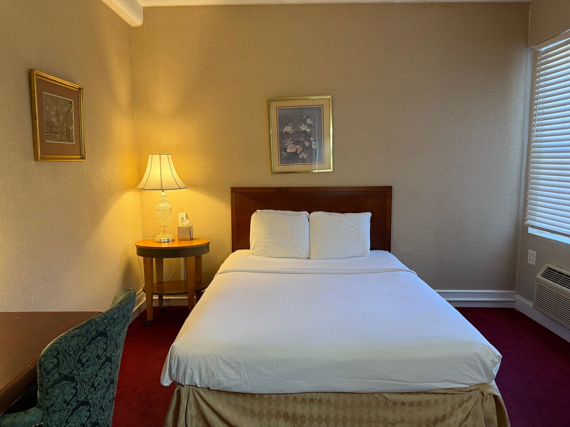 Hotel room with a bed, nightstand with lamp, artwork, and a window with blinds.