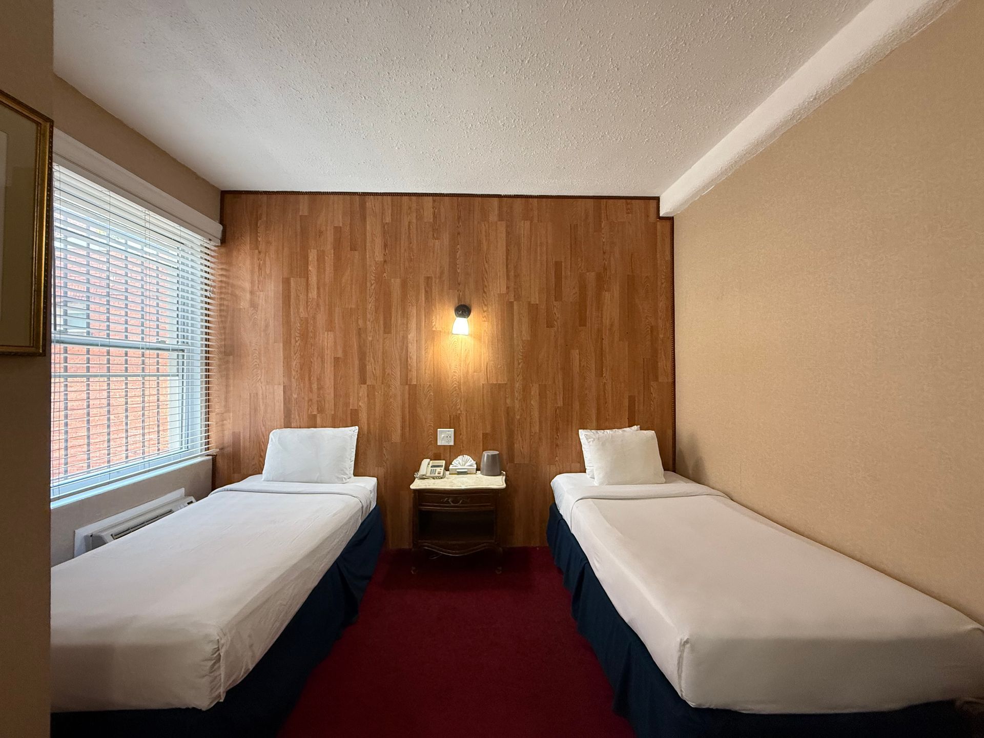 Two twin beds in a hotel room with wood paneling, a small table, and a window with blinds.