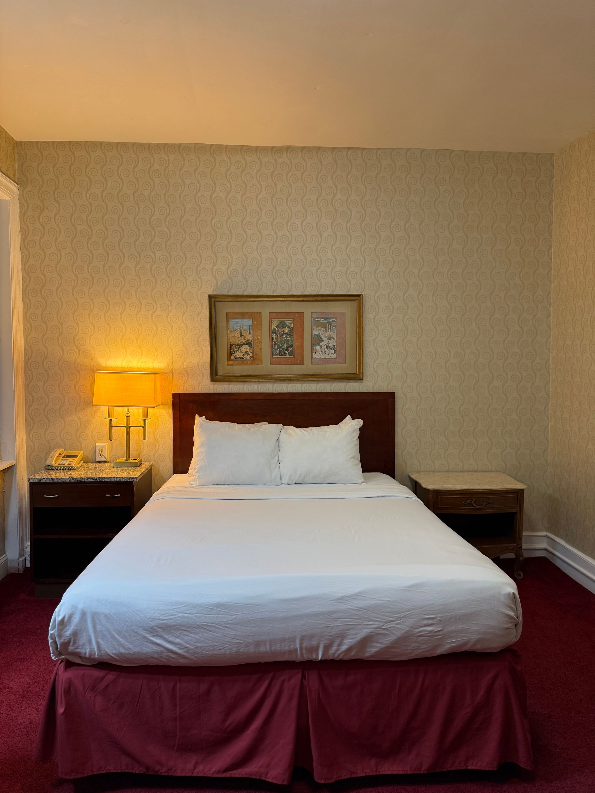 Hotel room with a bed, nightstands, lamps, artwork, and patterned wallpaper. Red carpet and bed skirt.