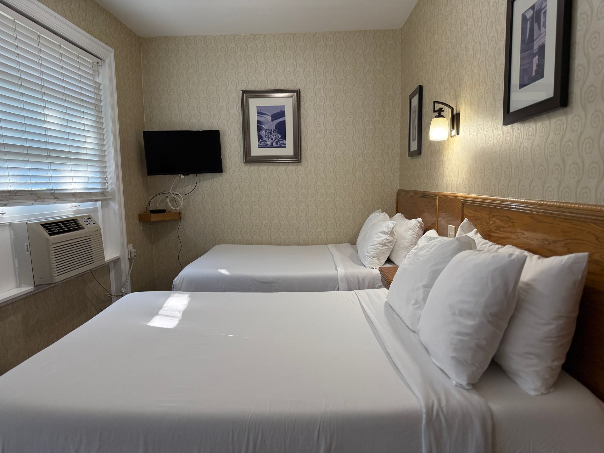 Two beds in a hotel room with white linens. Window with blinds, TV on wall, art.