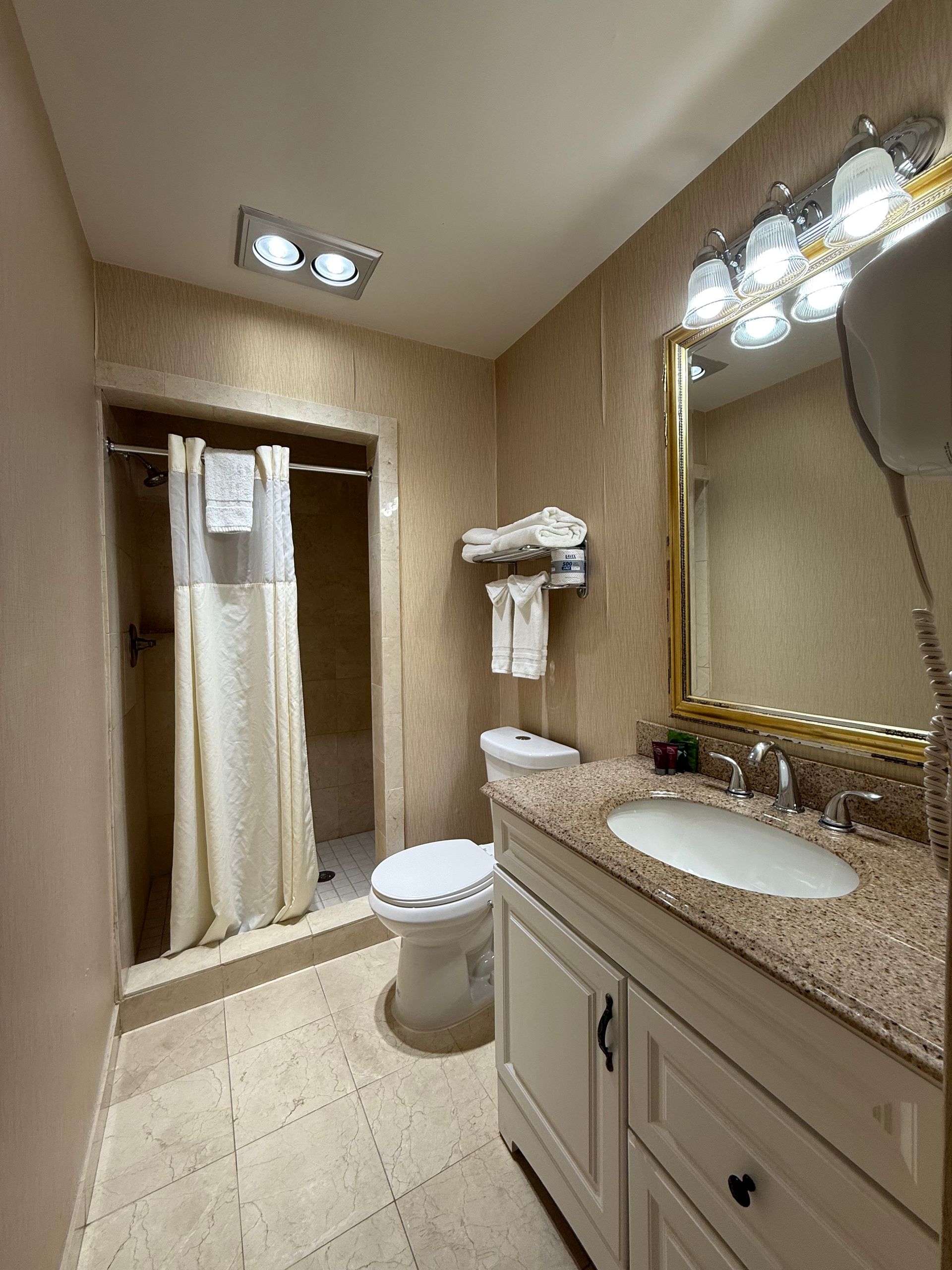 Bathroom with shower, toilet, sink, and mirror. Beige walls and light-colored tile floor.