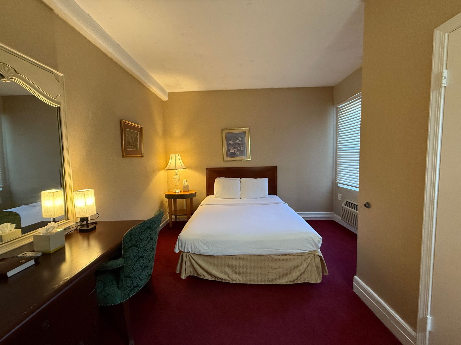 Hotel bedroom with a bed, desk, and window. Red carpet, beige walls, and a large mirror.