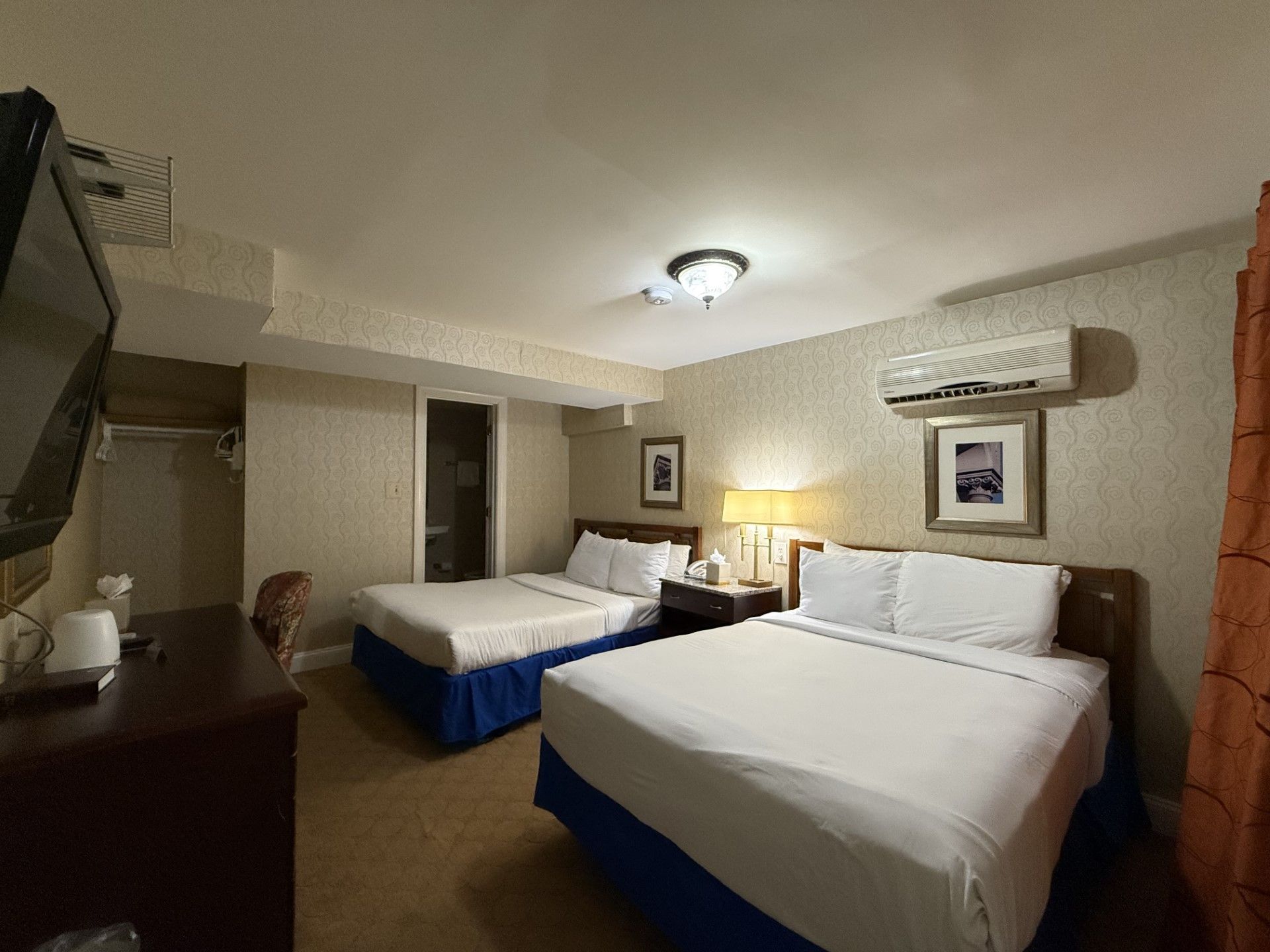A hotel room with two beds, a TV, and a dresser. The walls are beige, and the beds have white linens.