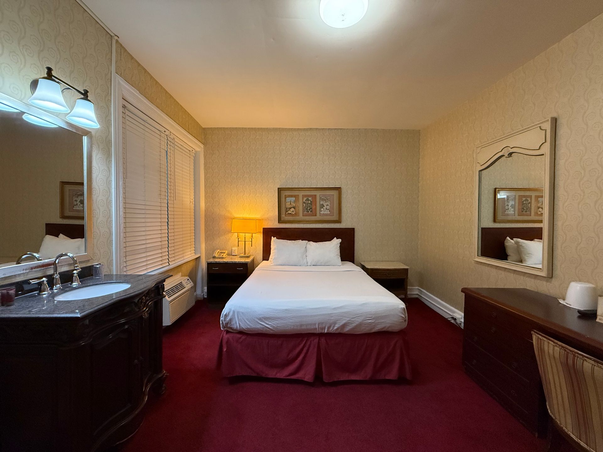 Hotel room with a bed, vanity, and mirror. Red carpet, beige walls, and warm lighting.