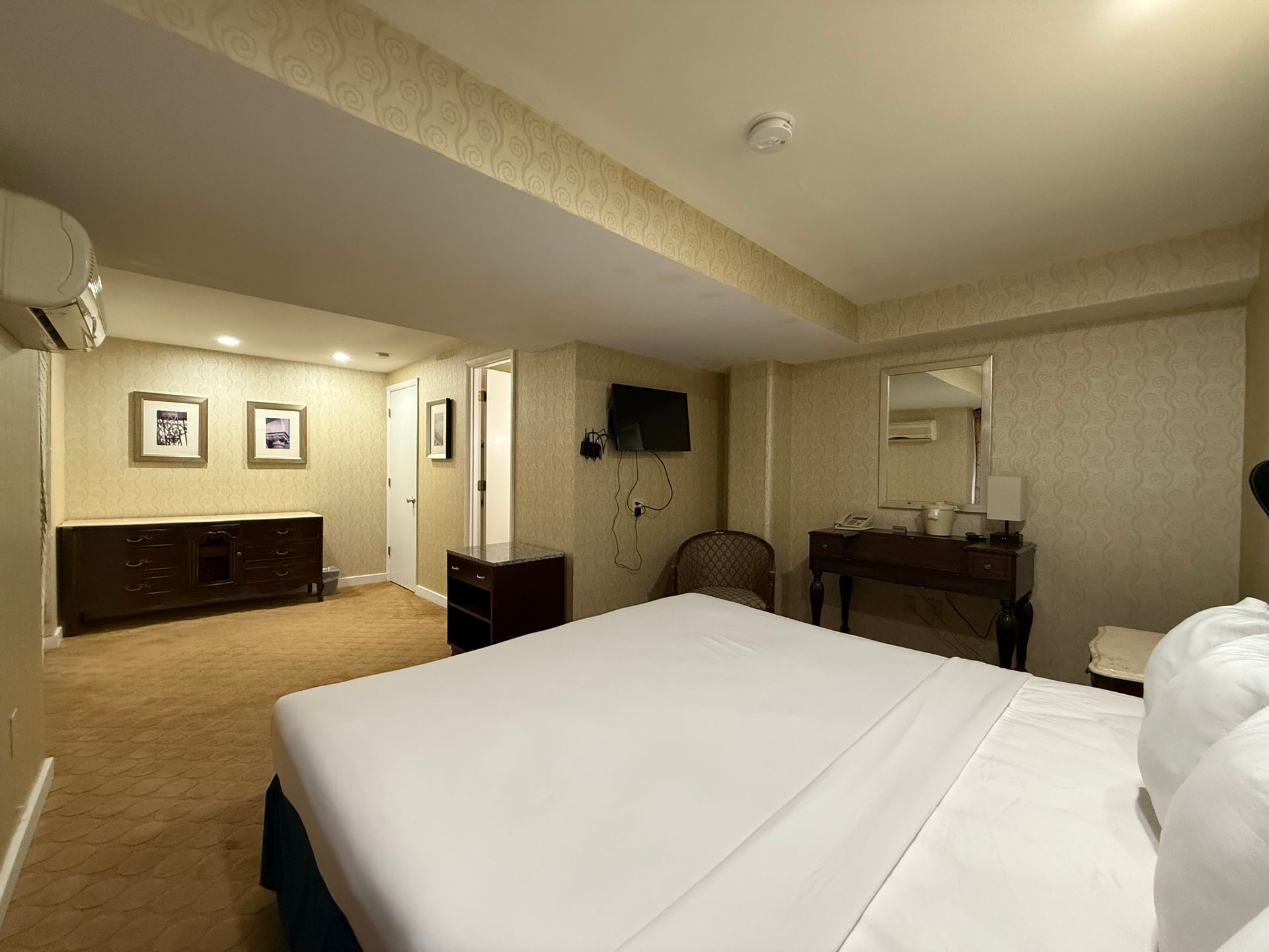 Bedroom with bed, dresser, TV, and vanity. Beige carpet and patterned wallpaper.