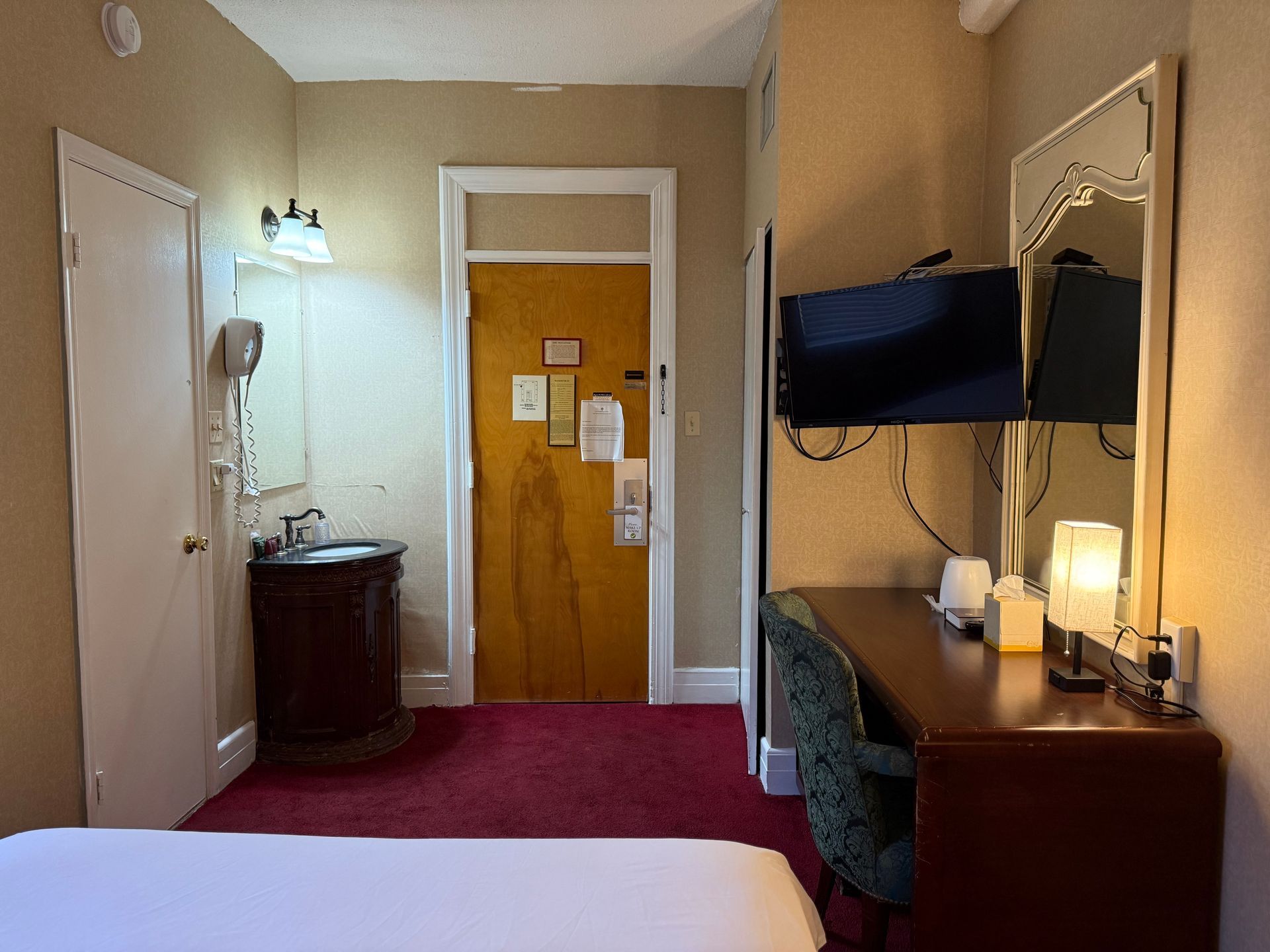 Hotel room with bed, desk, TV, sink, door, red carpet, and tan walls.