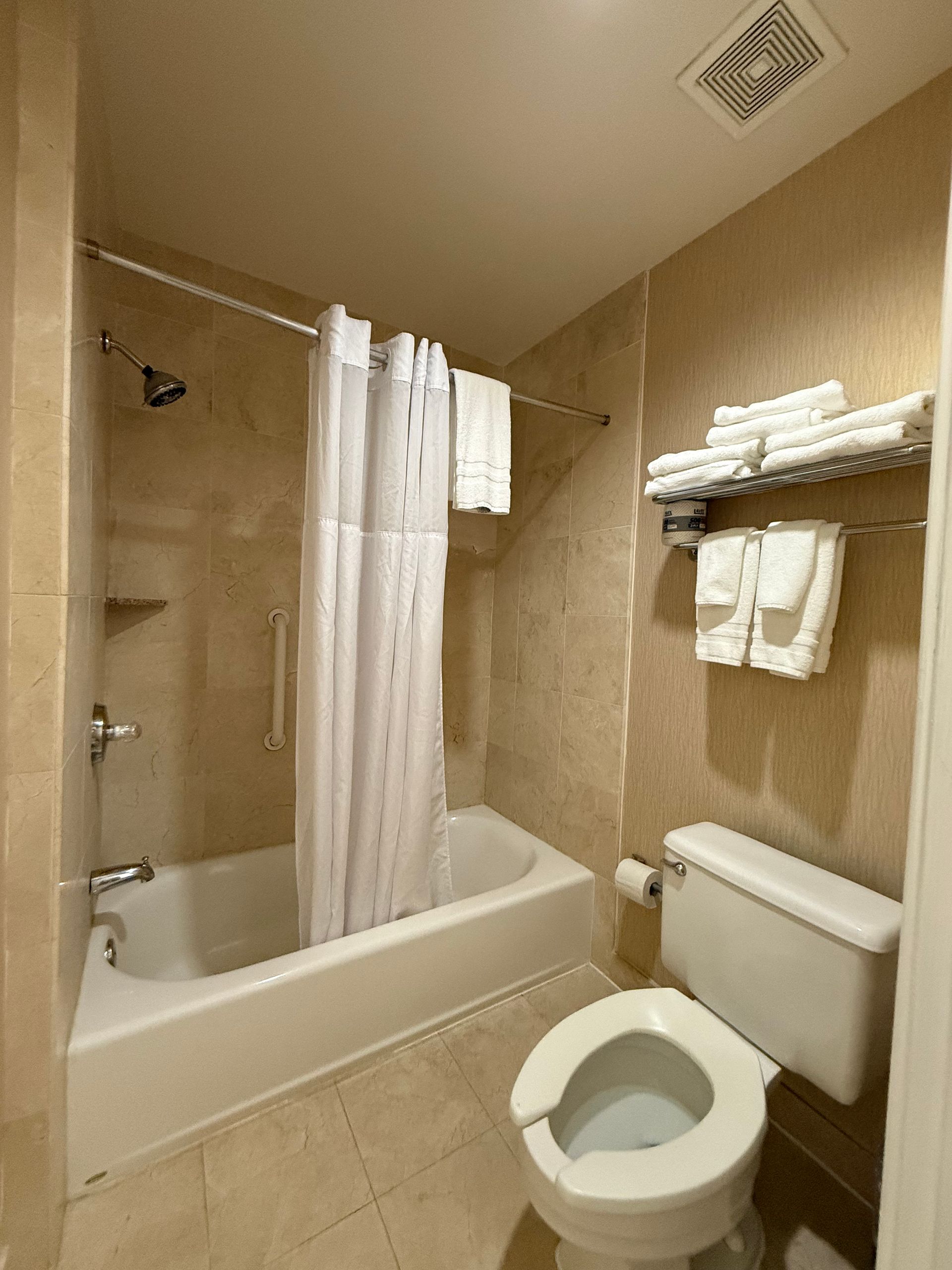 Bathroom with white tub, shower curtain, toilet, towels, and tan tile walls.