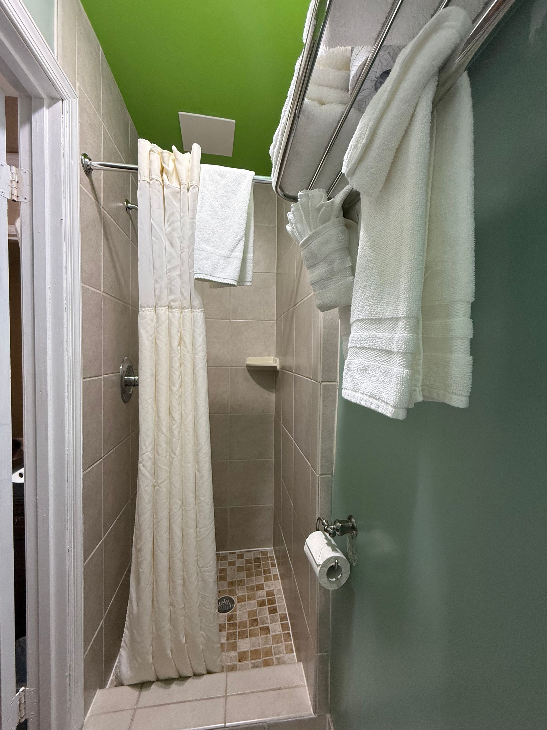Small shower with beige tiles, green ceiling and wall, white towels hanging, and shower curtain.