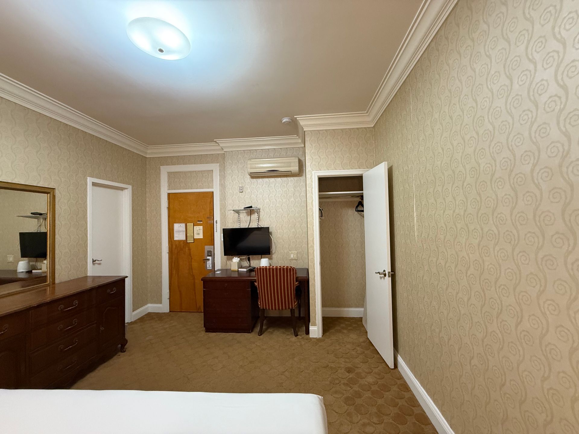 Hotel room interior with a dresser, desk, TV, closet, and patterned wallpaper.