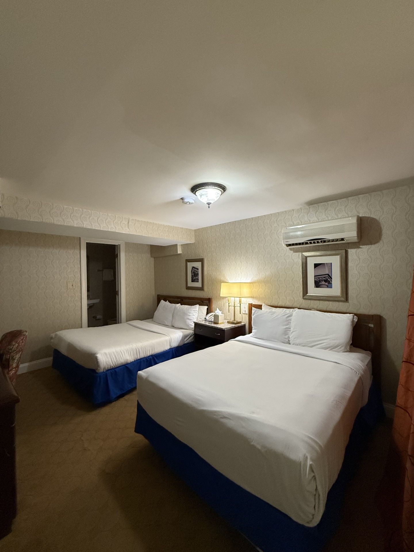 Hotel room with two double beds, white bedding, blue bedskirts, and a bathroom doorway.