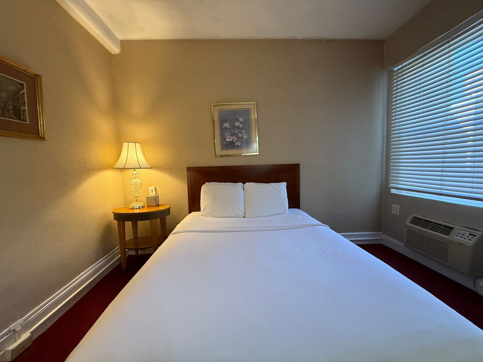 Hotel room with a made bed, nightstand with lamp, and a window with blinds.