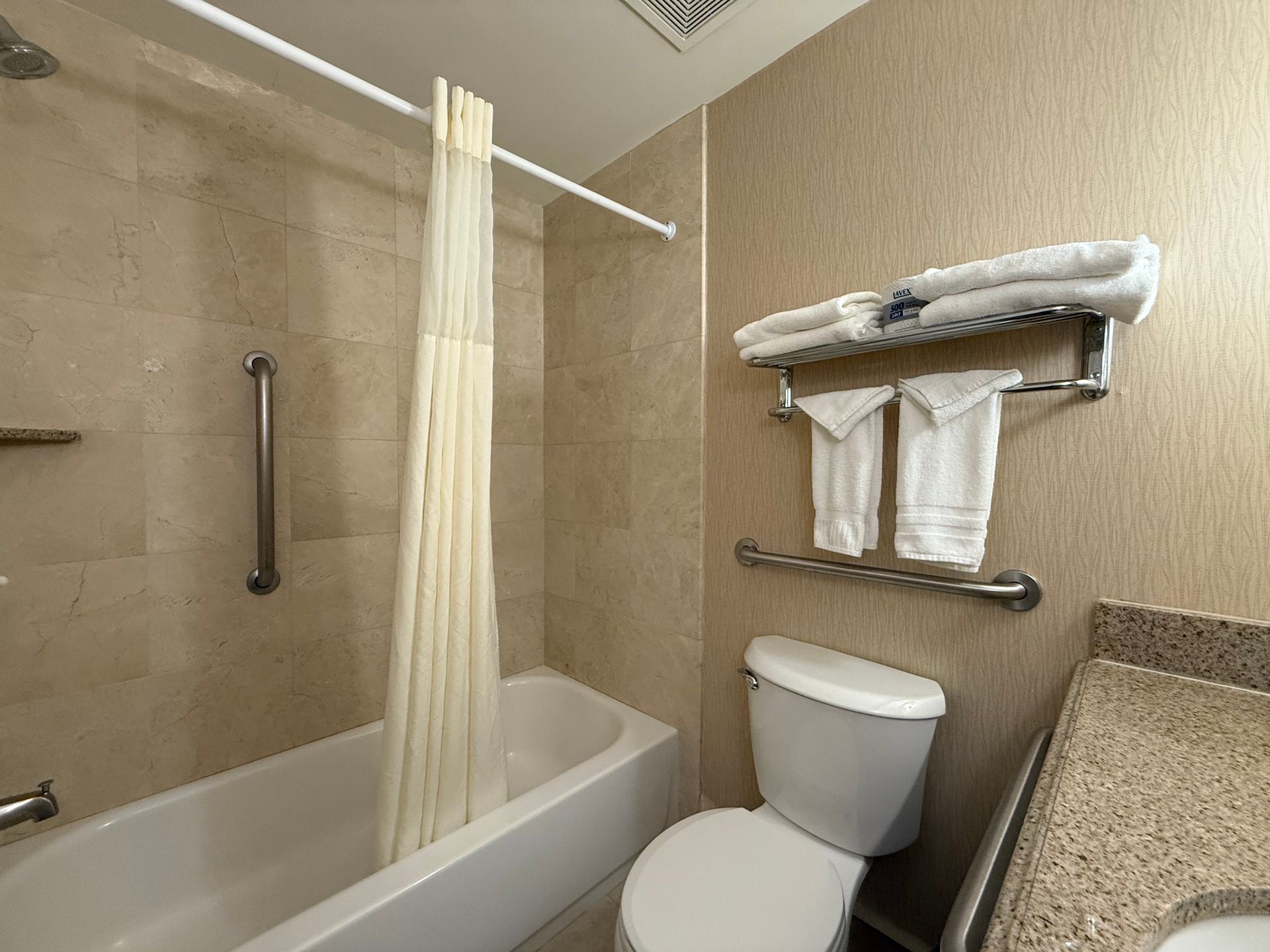 Bathroom with tub, toilet, and sink. White shower curtain, beige tile, and grab bars for accessibility.