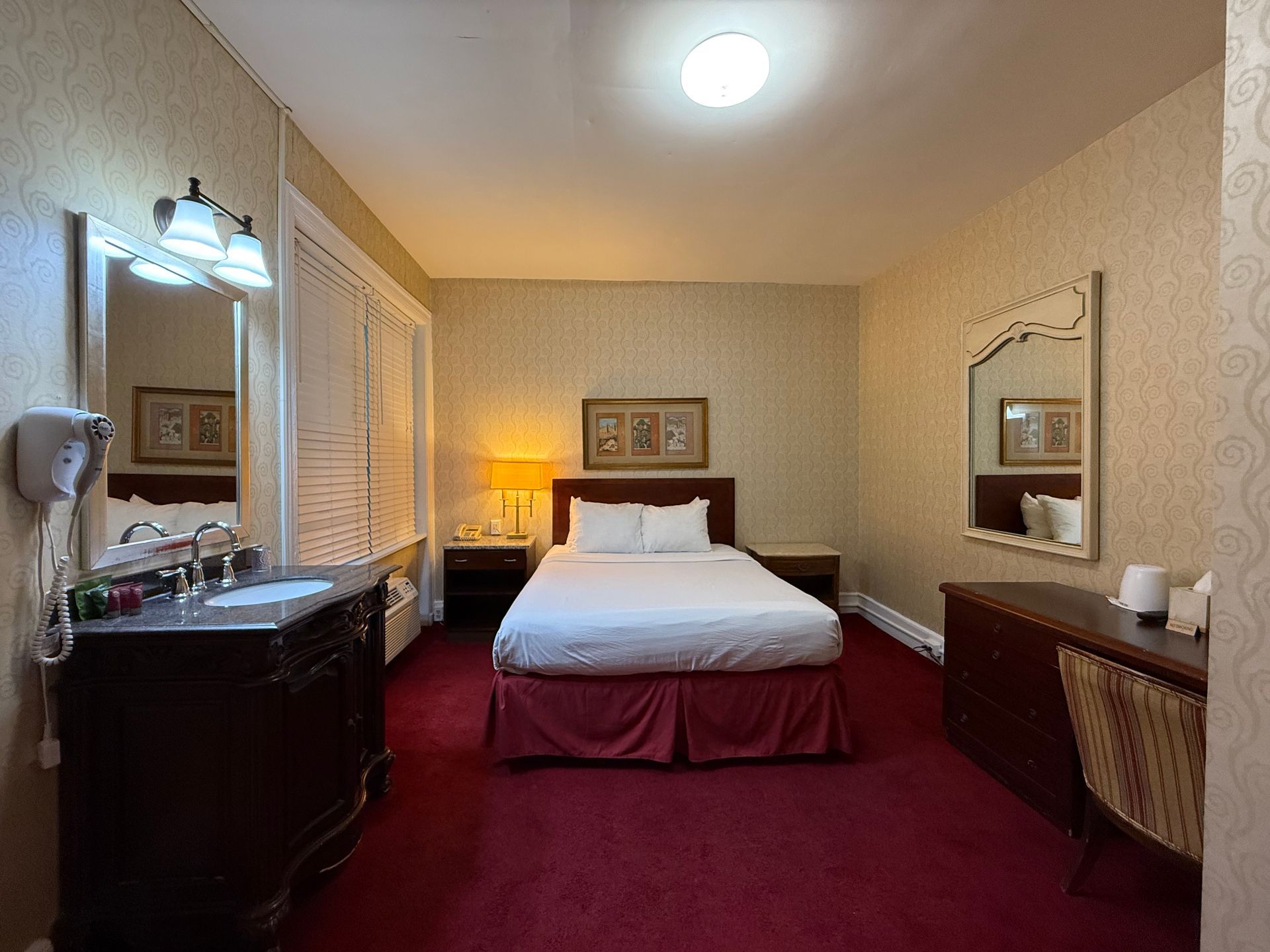 Hotel room with a bed, vanity, and desk on a red carpet. Beige walls and a bright ceiling light.