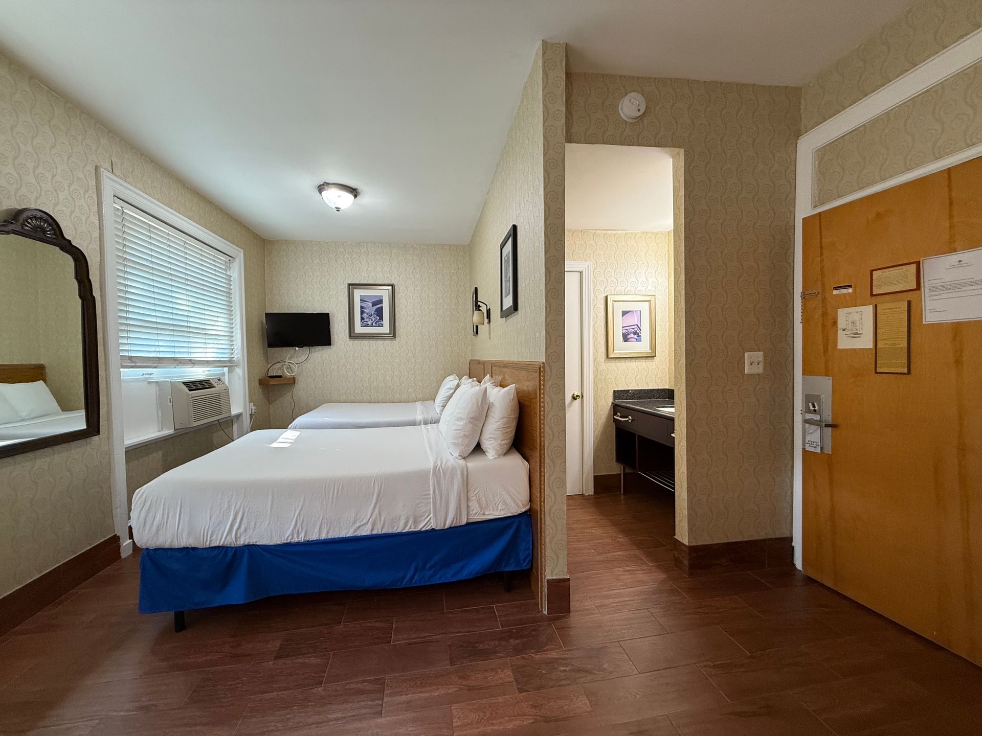 Hotel room with two beds, doorway to a bathroom, and a large mirror. Brown floor and tan walls.