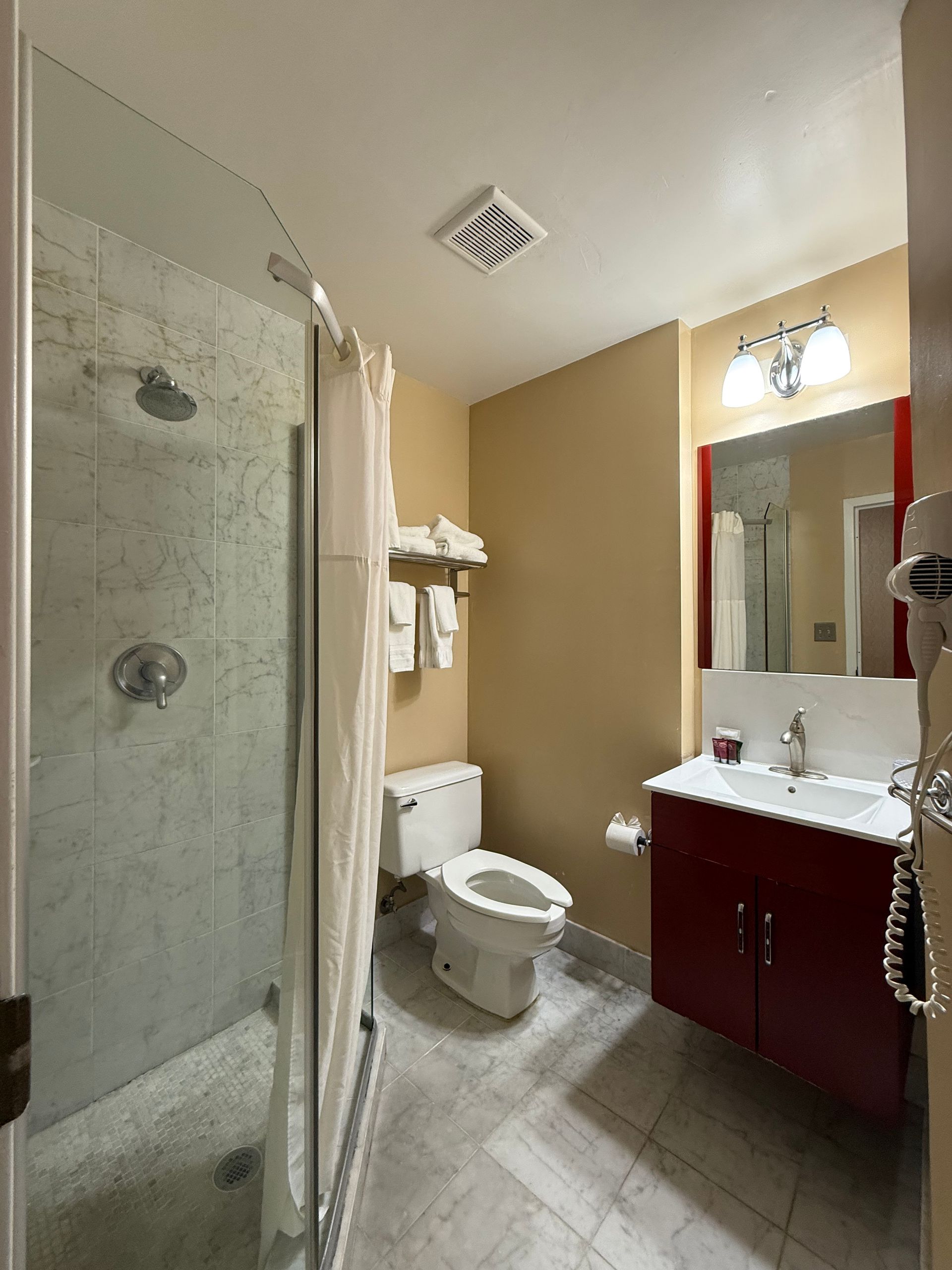 Bathroom with a shower, toilet, and red vanity. White shower curtain and beige walls.