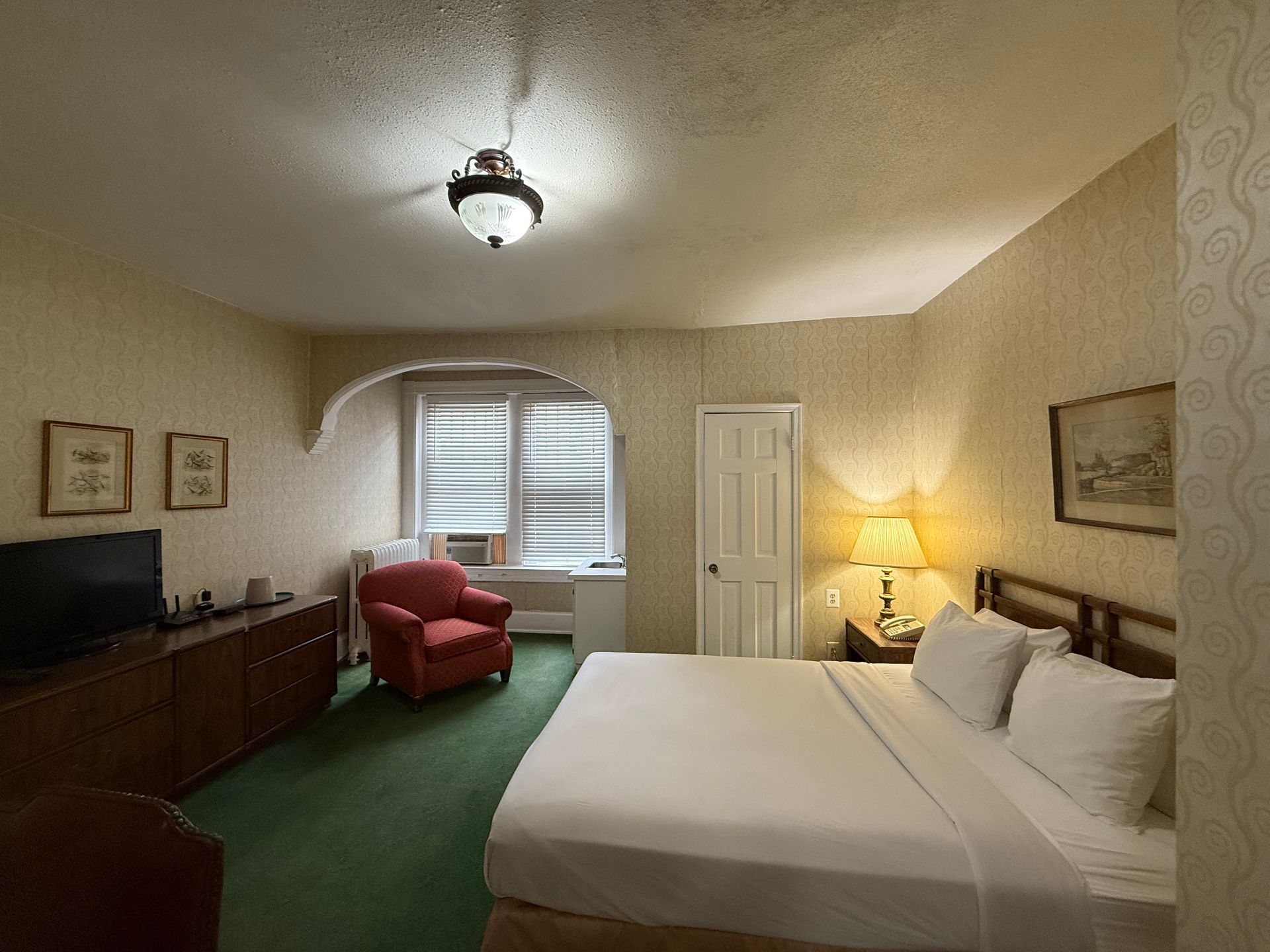 Hotel room with bed, chair, TV, and lamp; green carpet, light-colored walls.