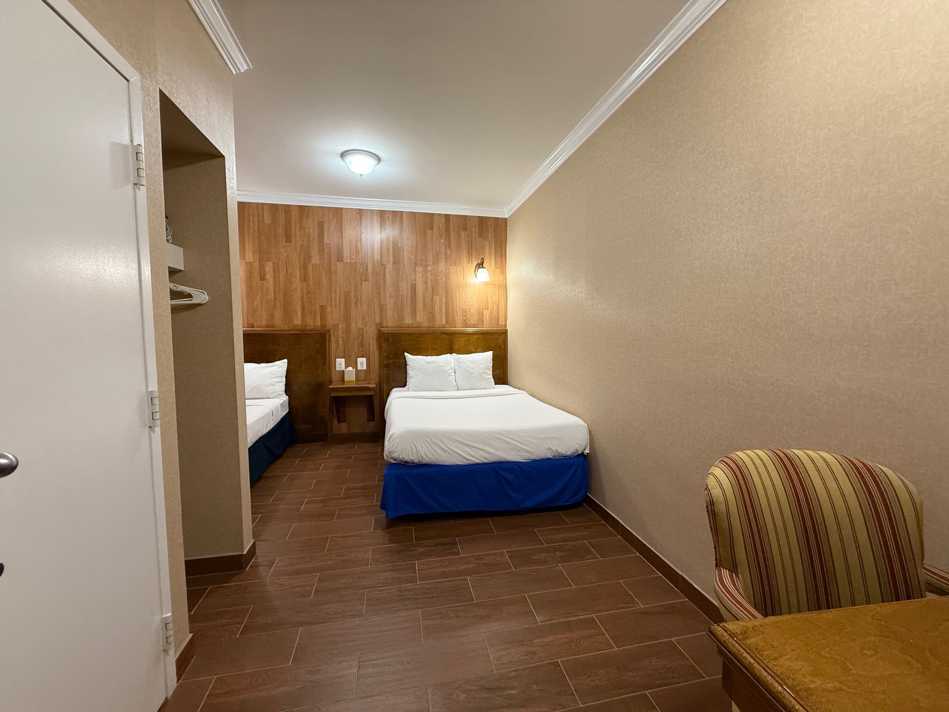 Hotel room with two twin beds, wood-paneled wall, blue bedskirts, chair, and brown tile floor.