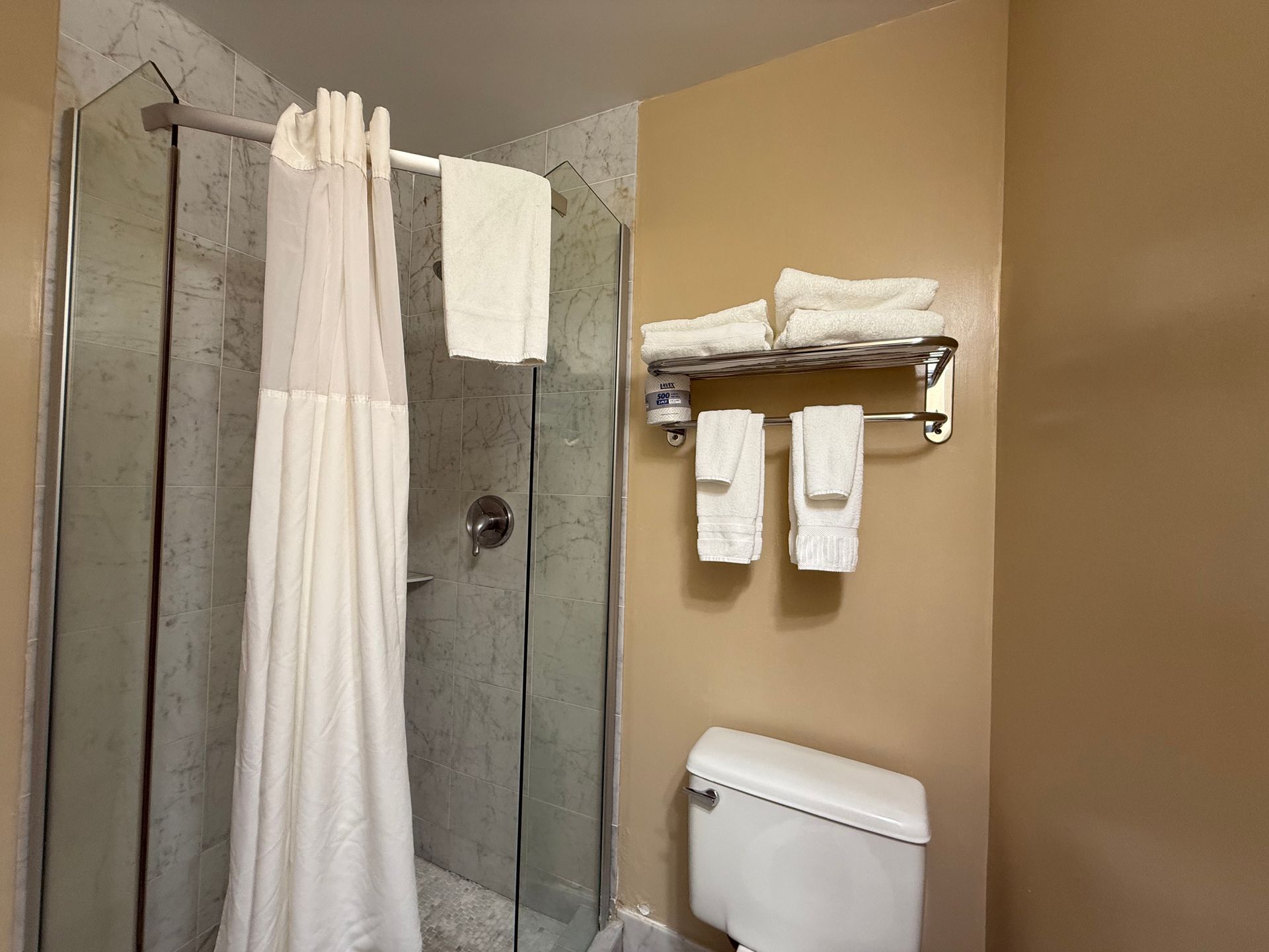 Bathroom with shower, toilet, and towel rack. Off-white towels and beige walls.