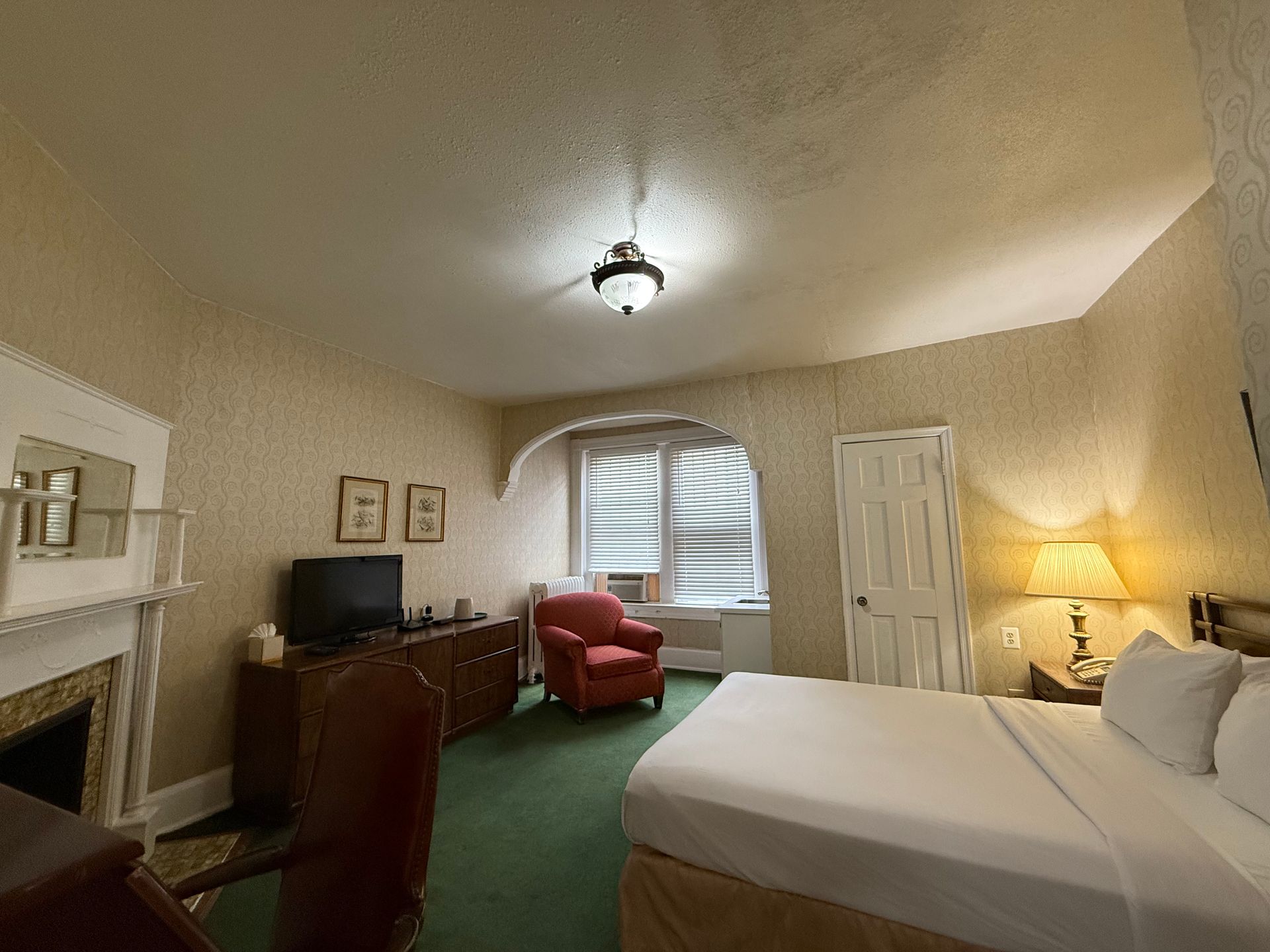 Hotel room interior: bed, fireplace, armchair, tv, desk, and arched window with blinds.