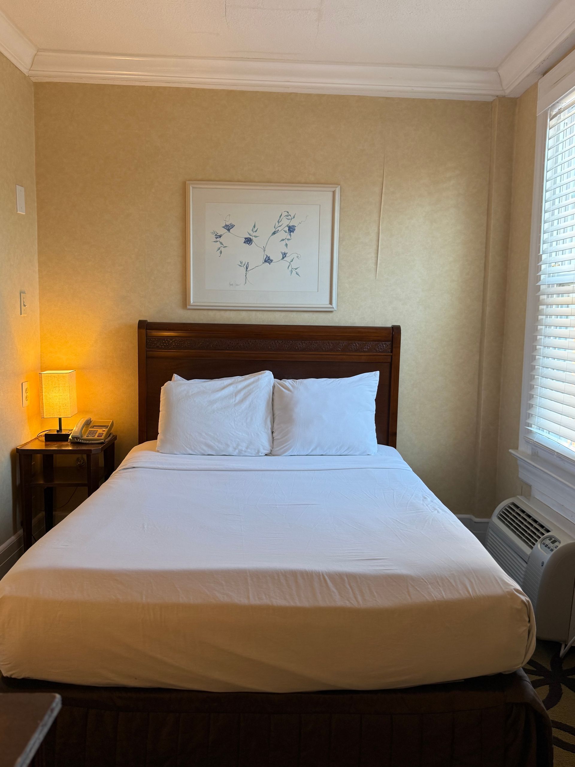 Bedroom with made bed, nightstand with lamp, and window with blinds.