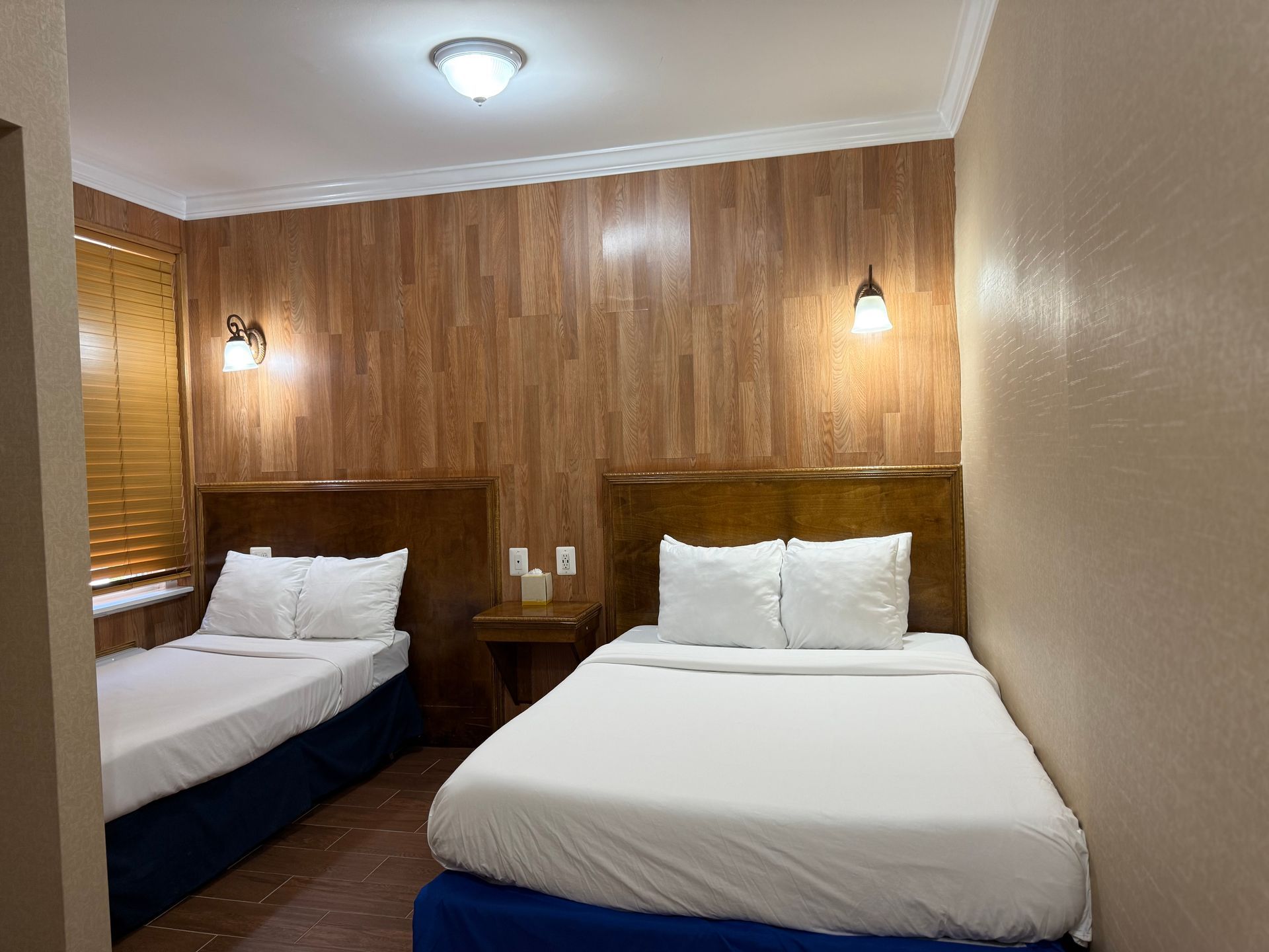 A hotel room with two beds, wood-paneled wall, overhead light, bedside lamps, and a closed window with blinds.