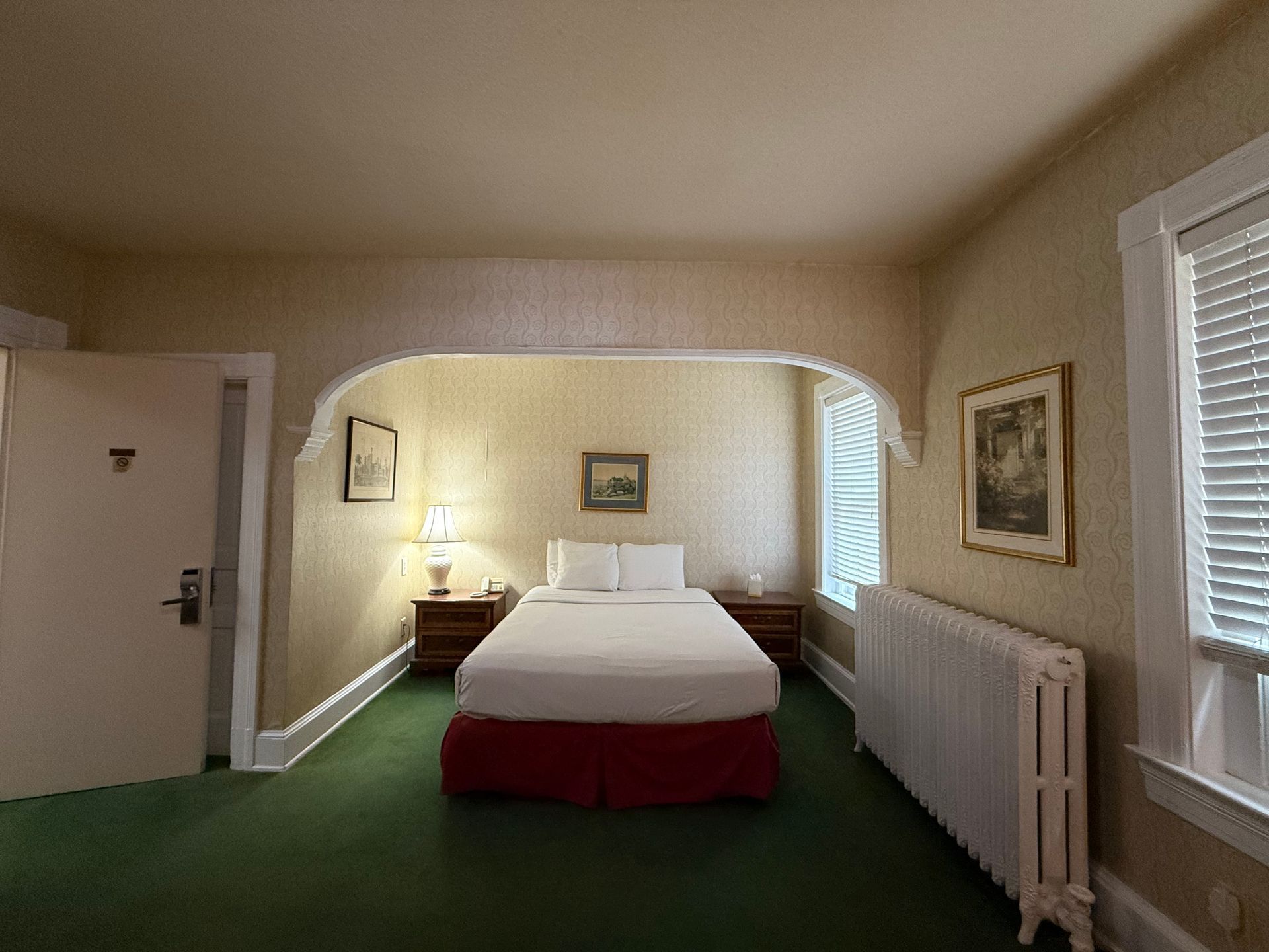 Hotel room with a twin bed, art, and radiator, in a green-carpeted room with beige walls.
