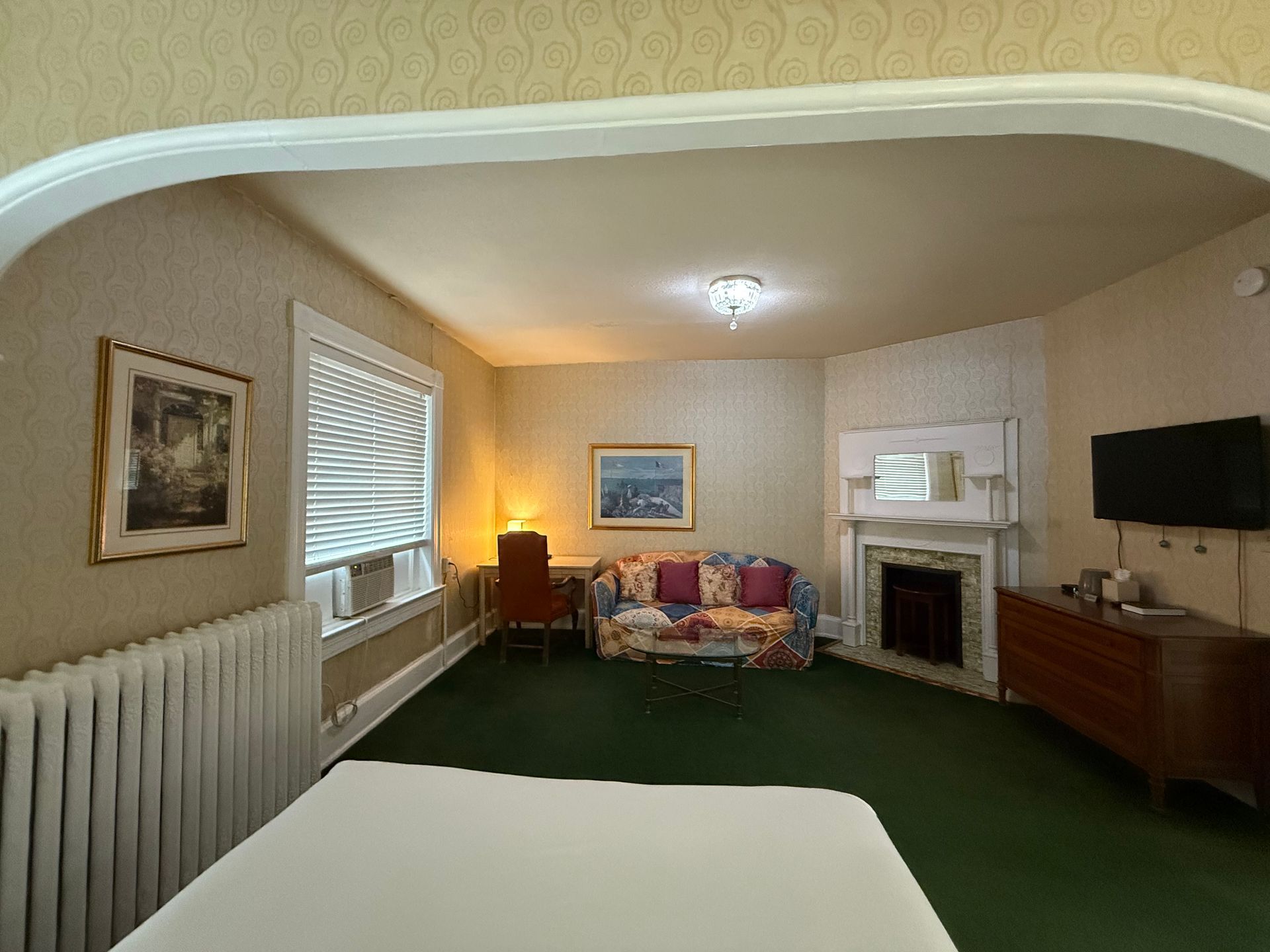 A hotel room with a green carpet, couch, fireplace, TV, and artwork. A window with closed blinds.