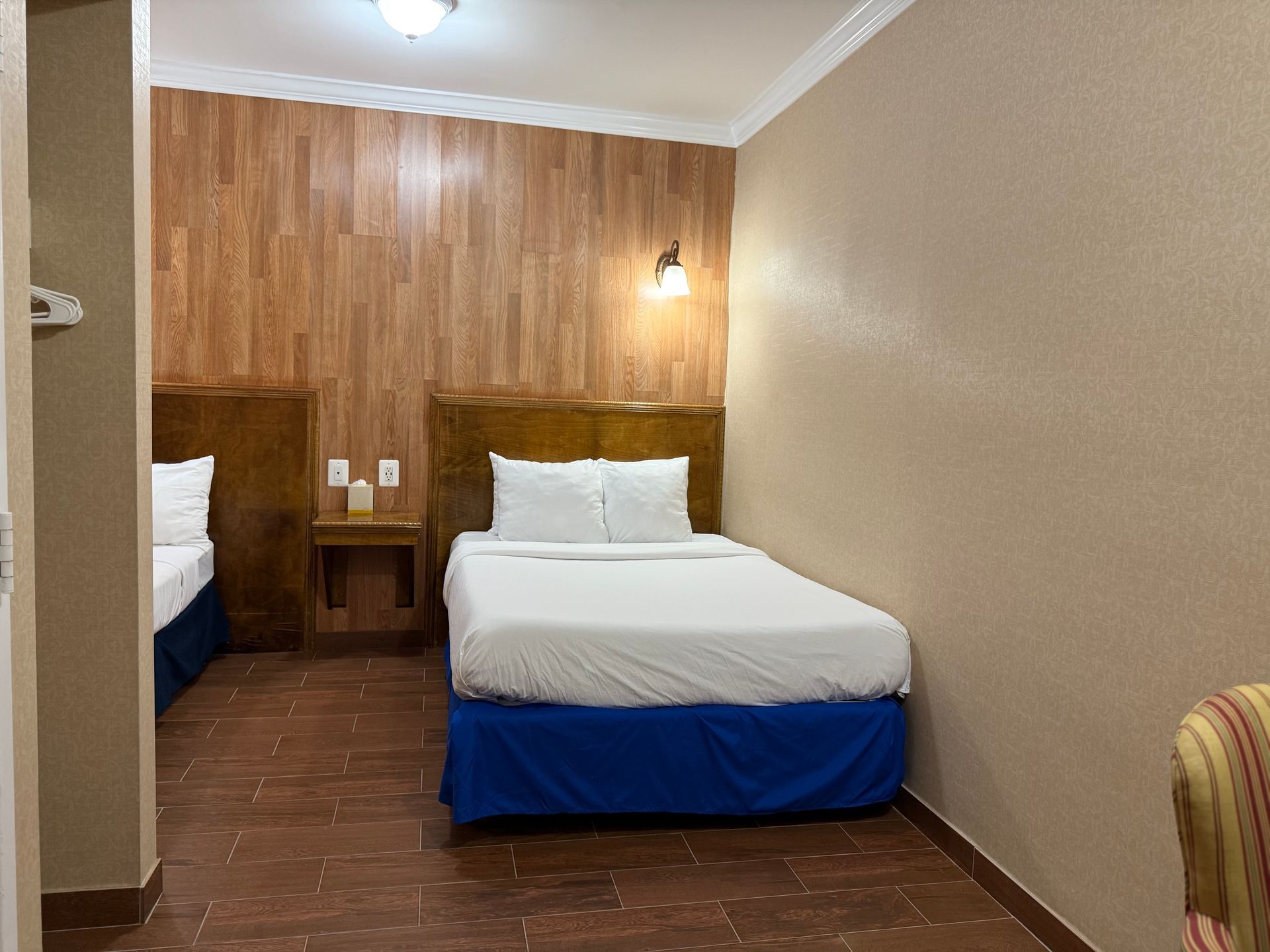 Hotel room with two twin beds, wood-paneled wall, nightstand, blue bed skirts, and brown tiled floor.
