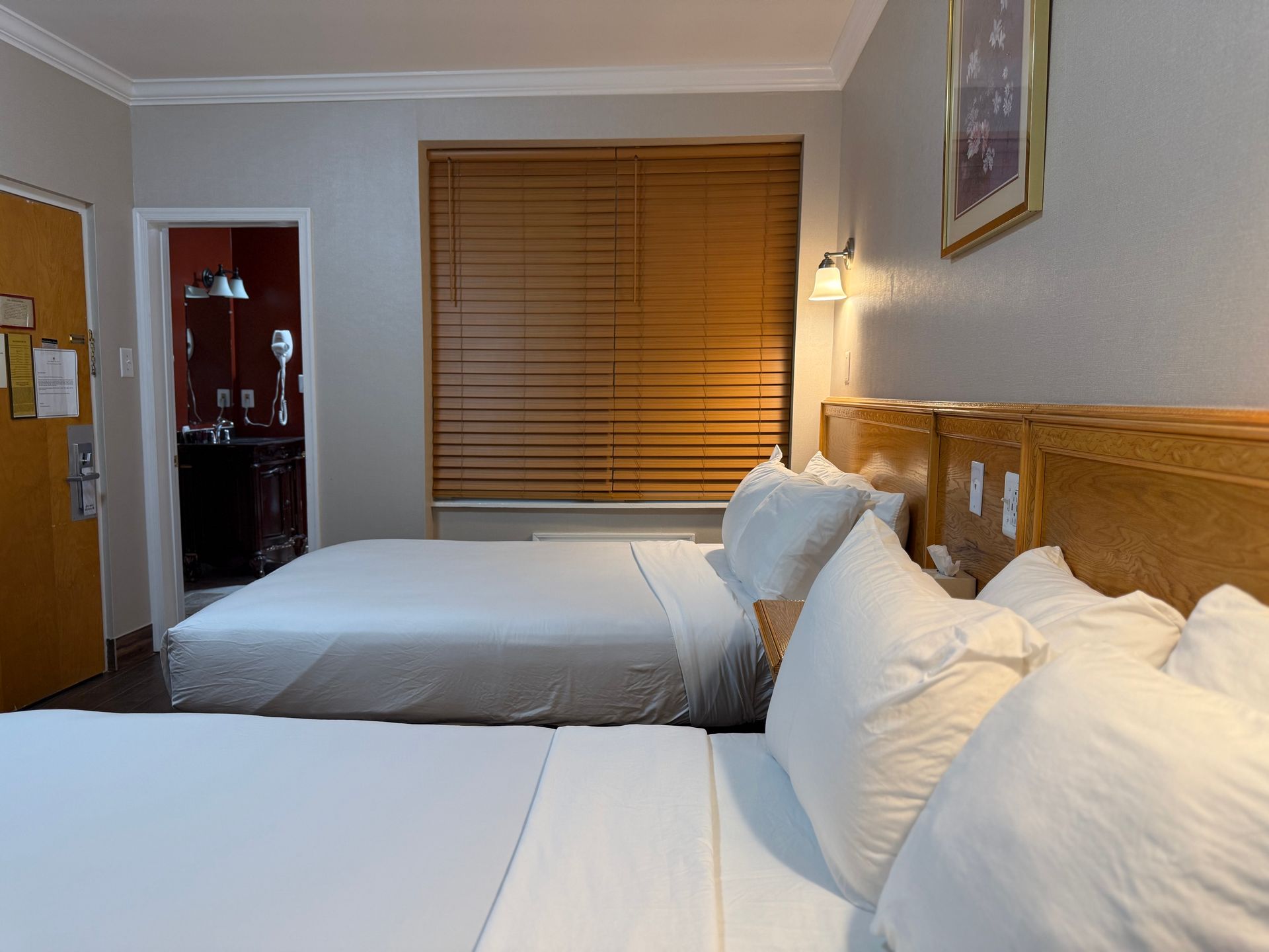 Two double beds in a hotel room with bathroom visible through doorway, window with blinds.