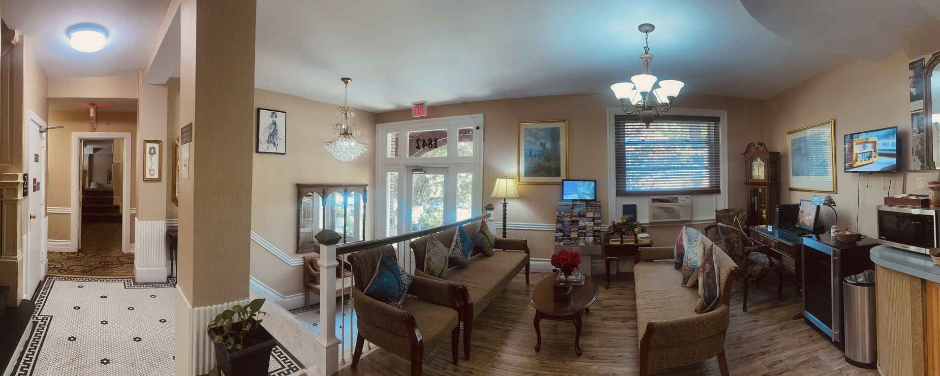 Interior of a building with a hallway and a living room. Sofas, tables, and artwork are visible.