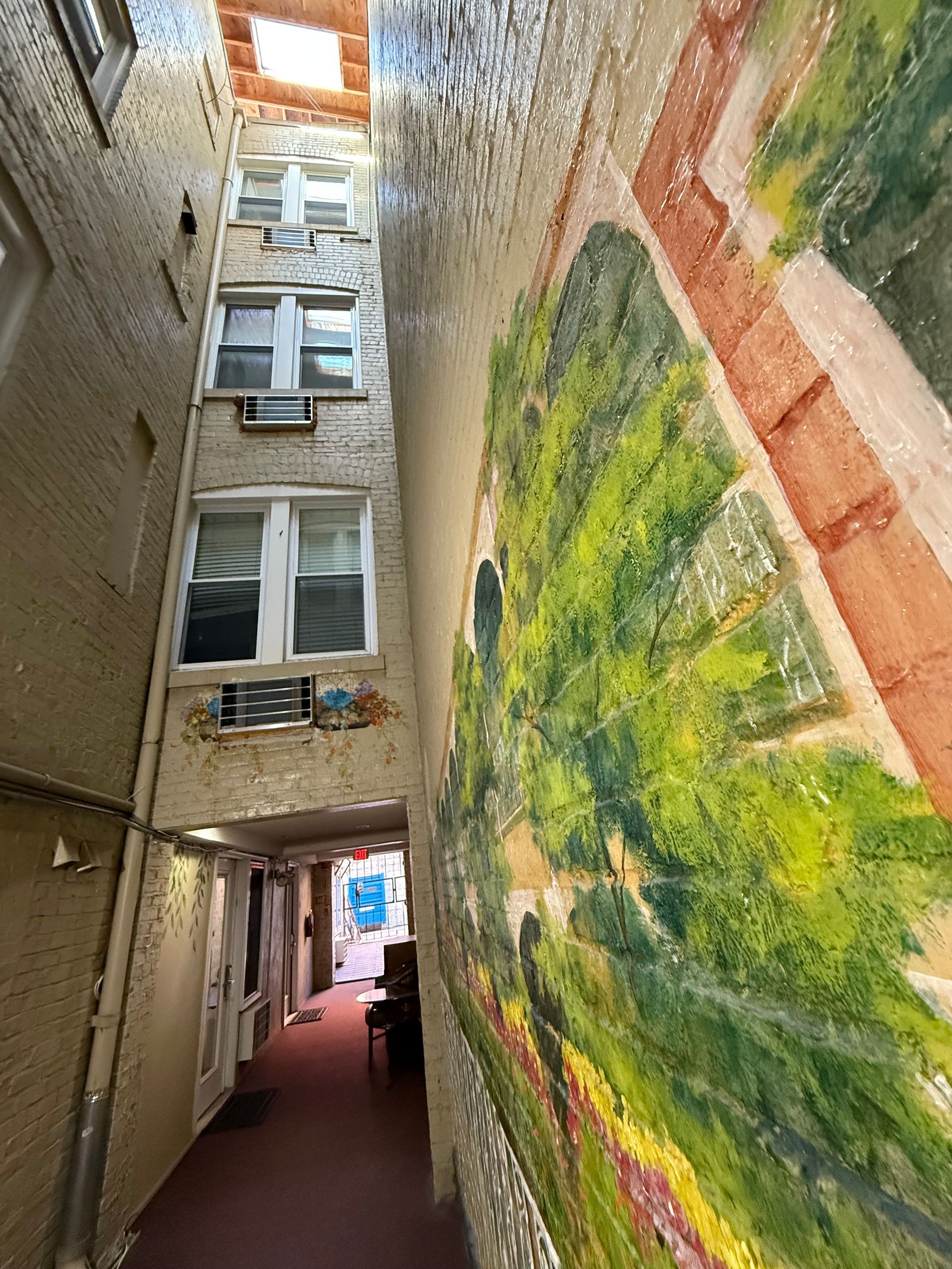 Narrow alleyway between buildings, one with mural of green landscape. Windows visible; sunlit at end.