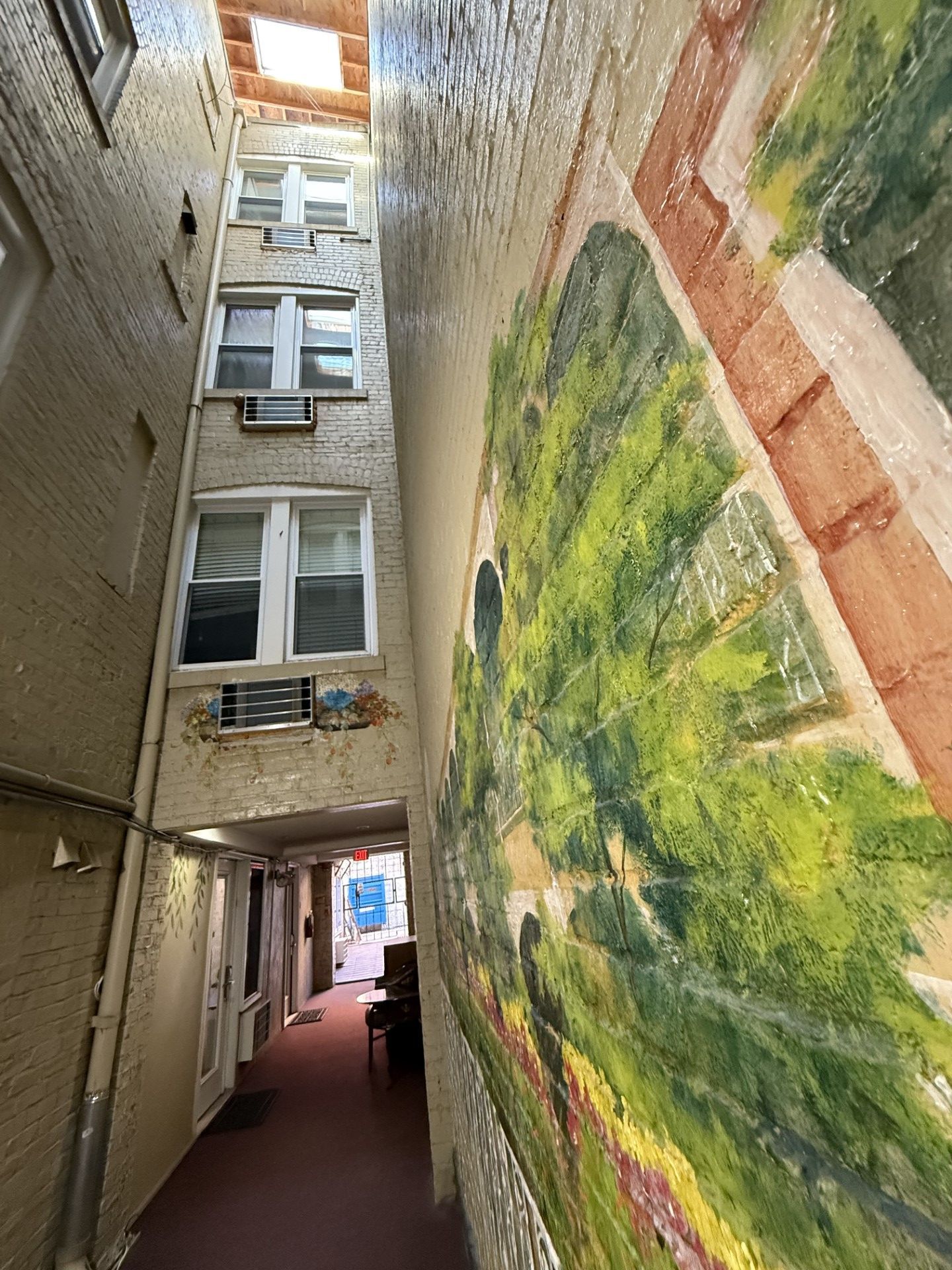 Narrow alleyway between brick buildings, one wall with a mural of a green landscape.