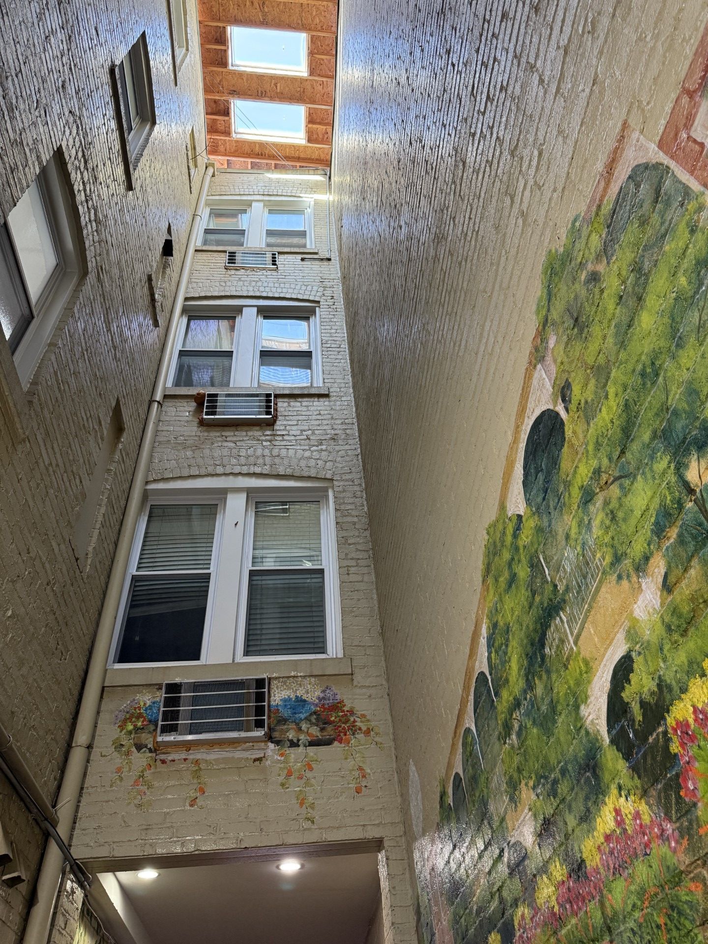 Narrow urban alleyway with windows on one side and a mural on the other, visible skylights at top.