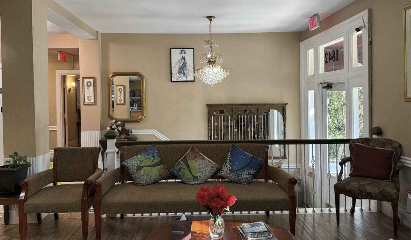 An elegant lobby with sofa, chairs, chandelier, mirror, artwork, and exit doors. Beige walls, wood trim.