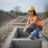Woman in a hard hat and safety vest squats on a concrete structure at a construction site, smiling.