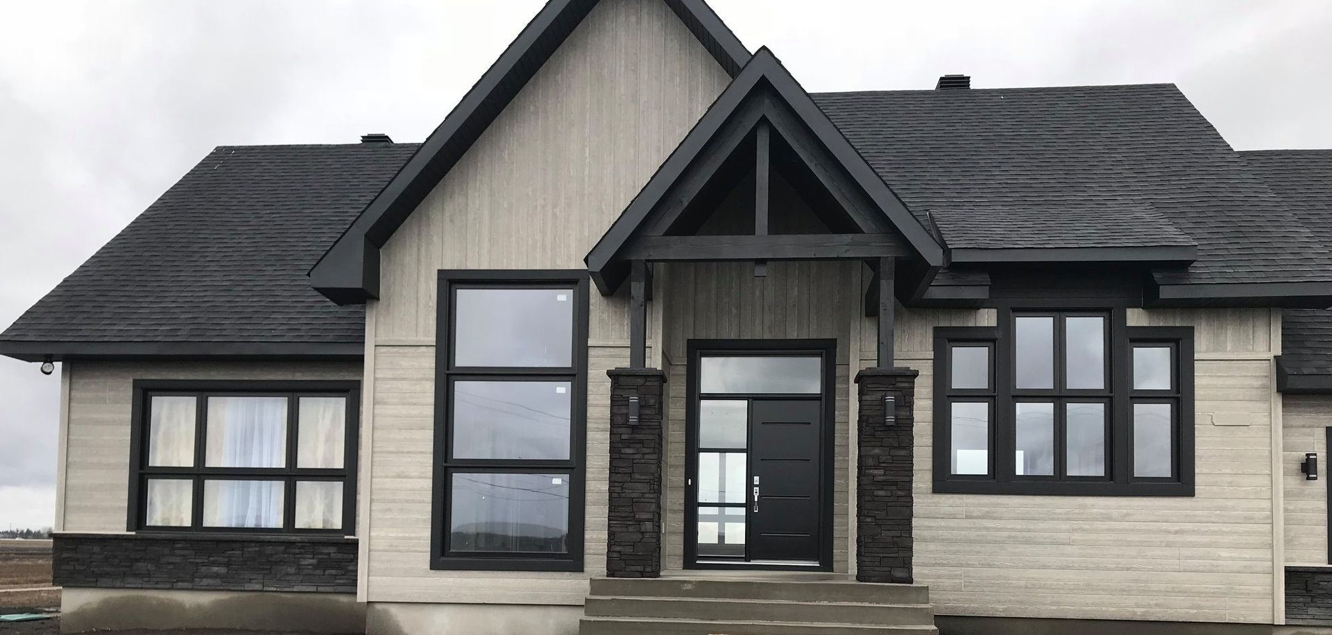 Maison moderne avec bardage gris, toit et moulures noirs. Grandes fenêtres et porte d'entrée sombre.