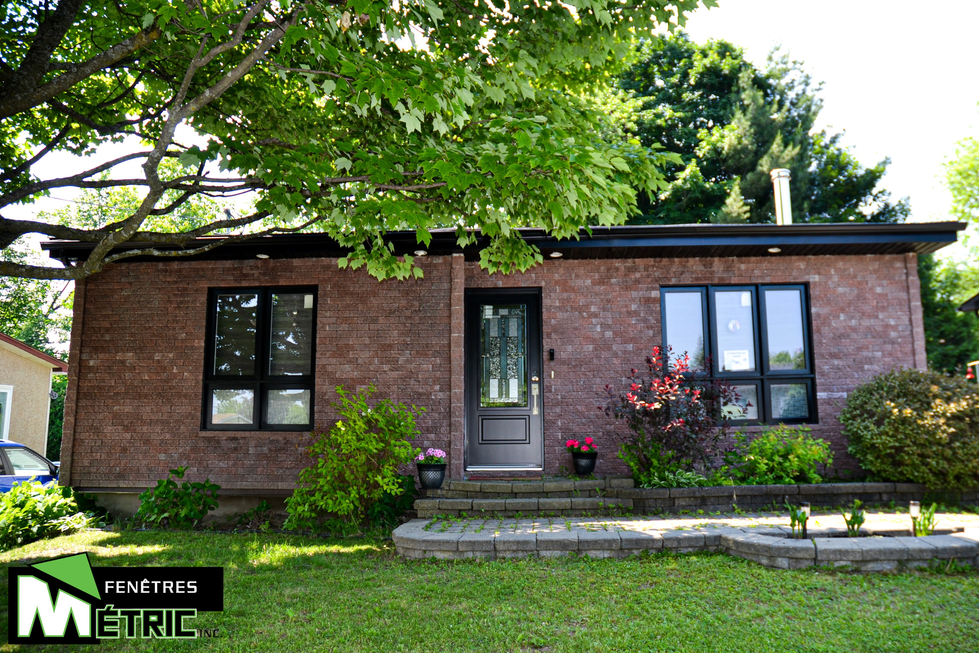 Bungalow en brique avec fenêtres et porte à cadre noir, cour avant avec verdure, sous un arbre feuillu.