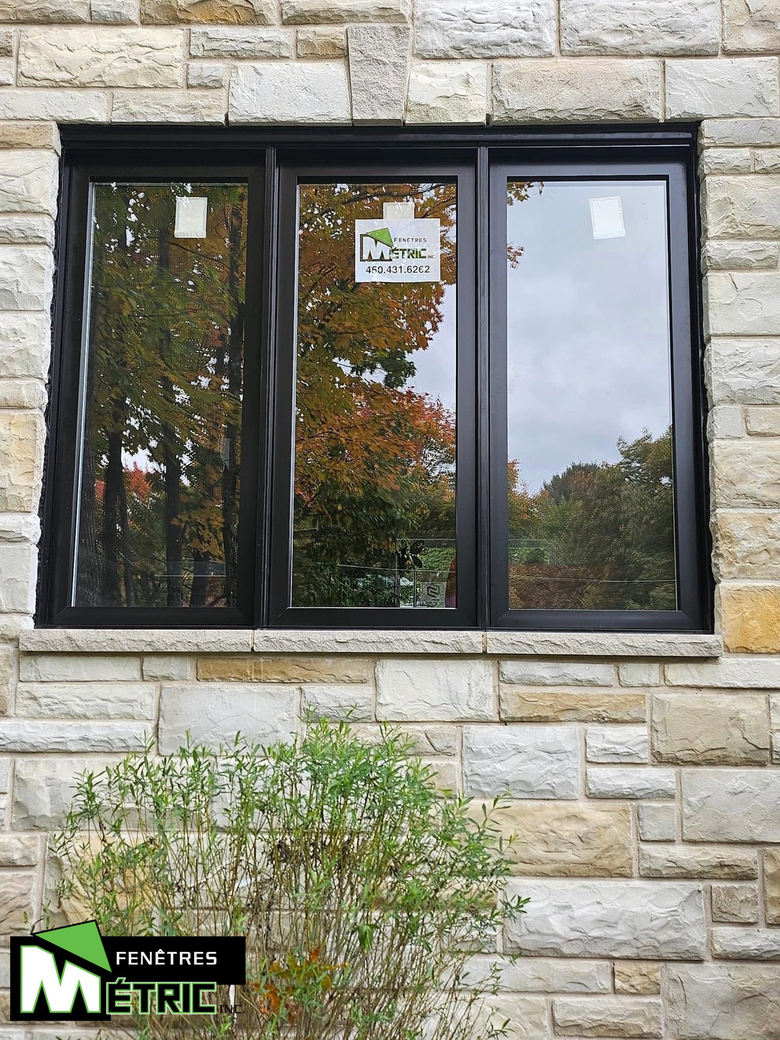 Fenêtre coulissante à cadre noir encastrée dans un mur en pierre, reflétant un arbre au feuillage d'automne.