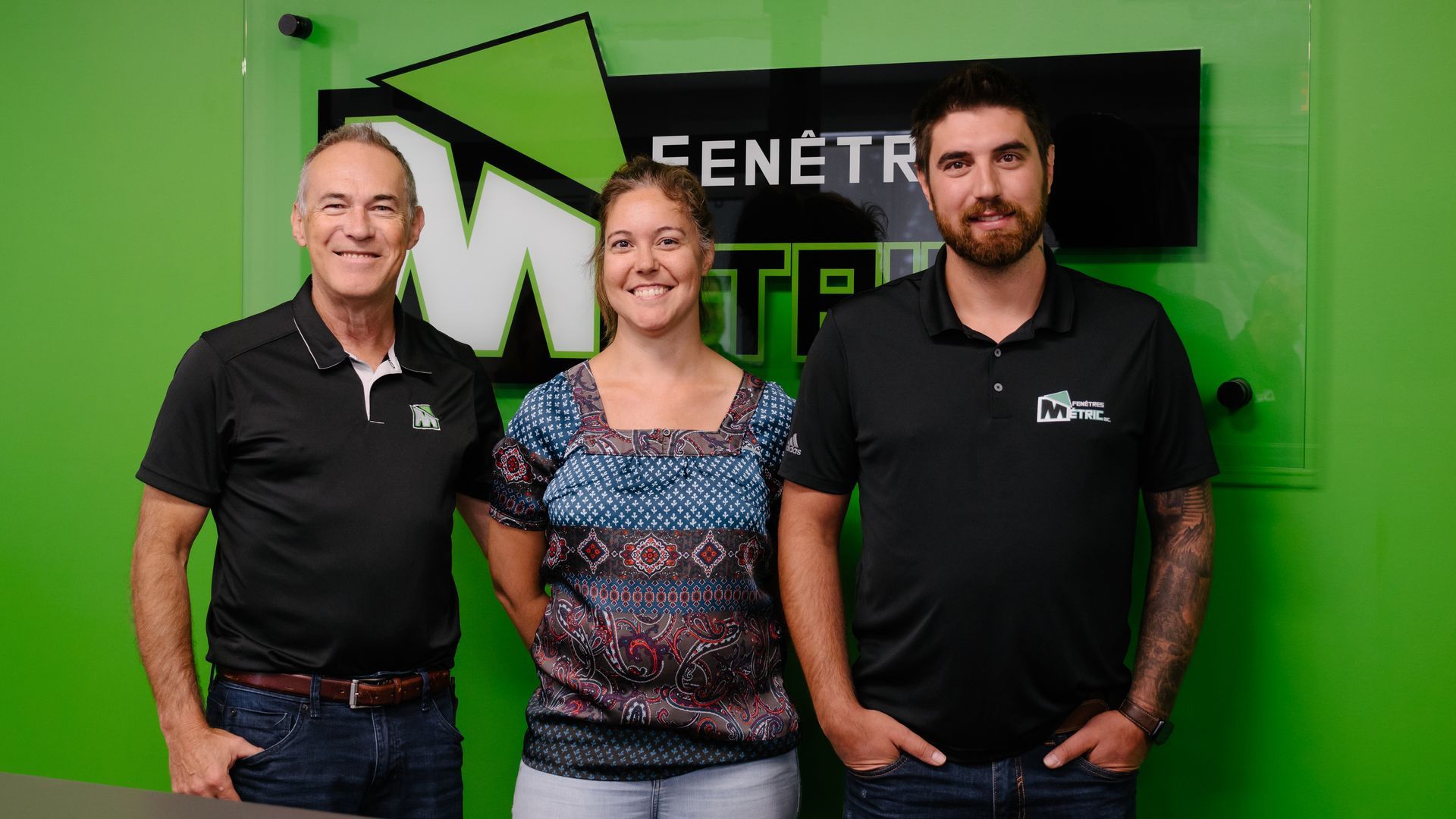 Trois personnes se tiennent devant un panneau vert. Un homme à gauche, une femme au milieu, un homme à droite. Tous sourient.