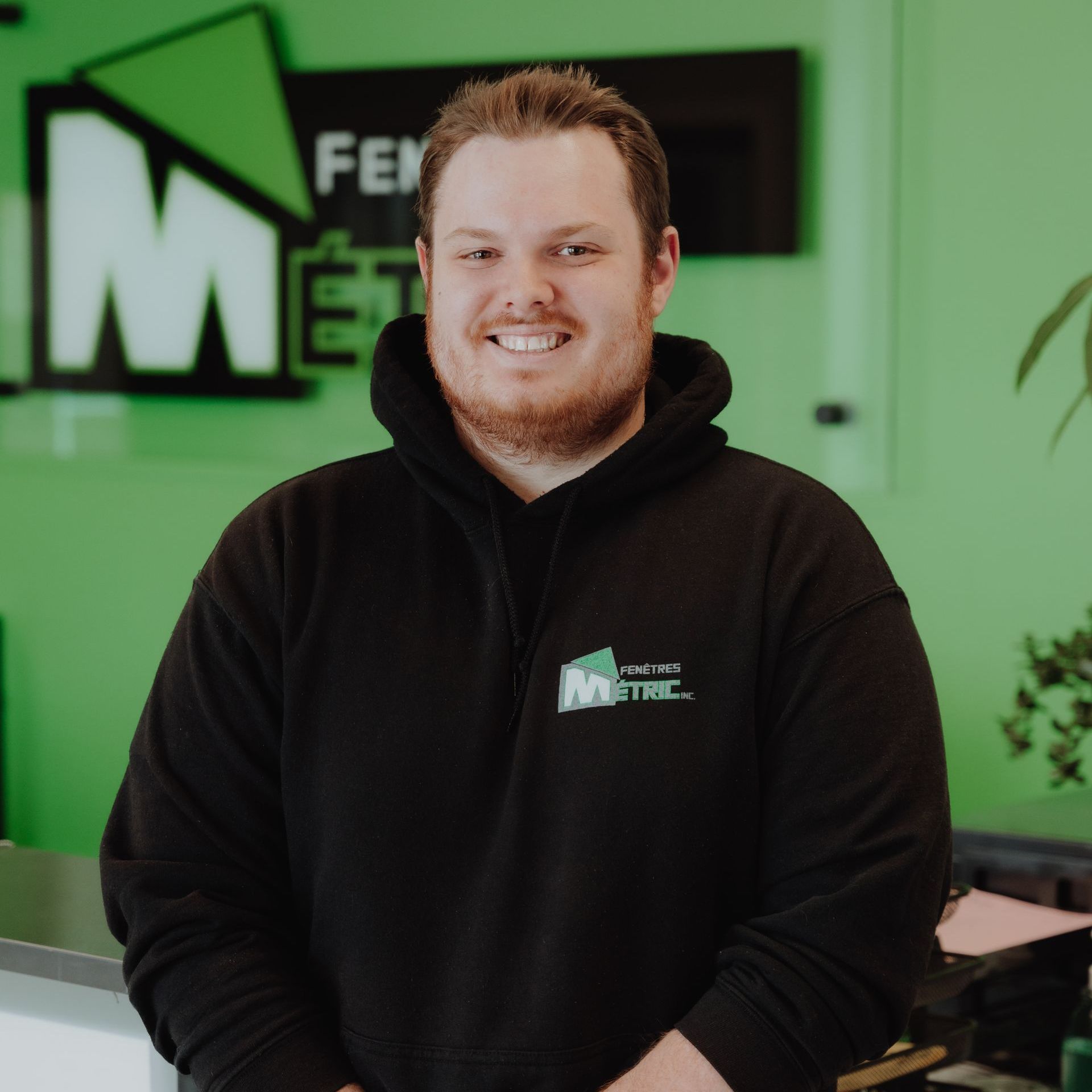 Un homme en sweat à capuche noir souriant devant un panneau vert ; prise de vue intérieure.