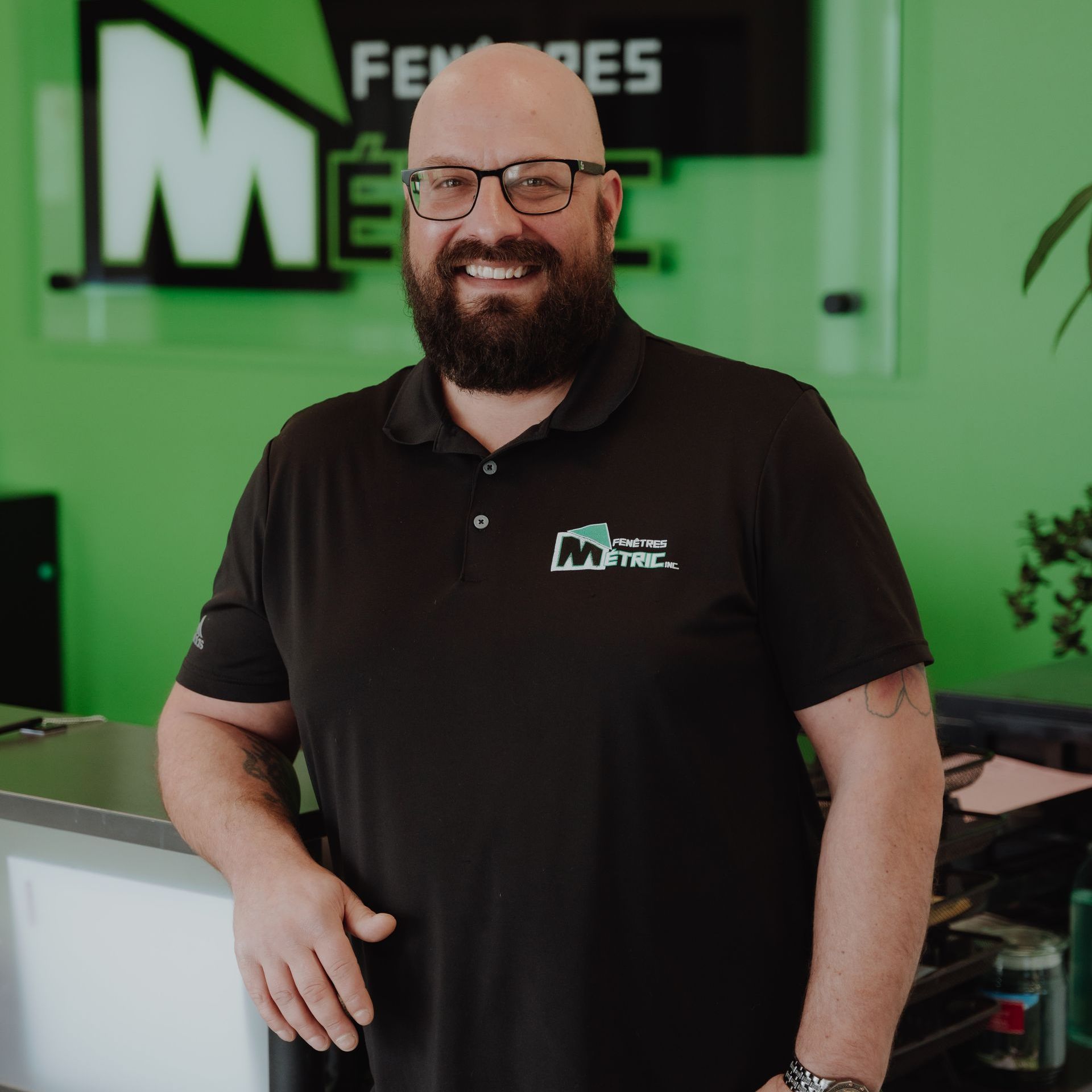 Un homme avec une barbe et des lunettes sourit, appuyé sur un comptoir dans un bureau avec un mur vert.