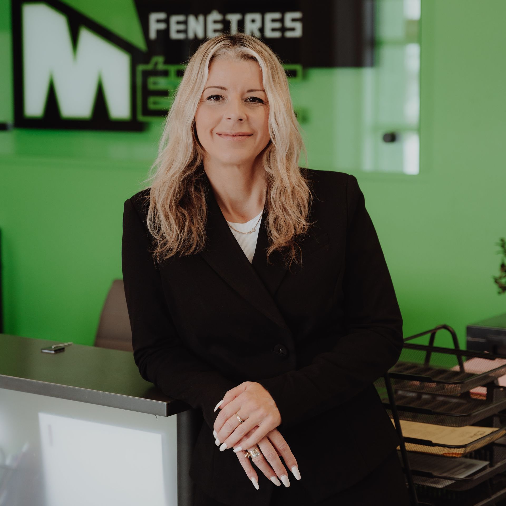 Femme en blazer noir appuyée sur un bureau, souriante, dans un bureau aux accents verts.