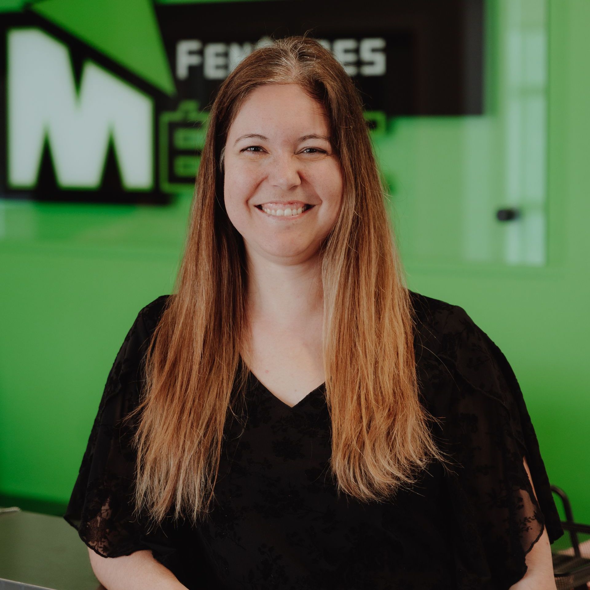 Une femme aux cheveux longs souriant à la caméra dans un bureau aux murs verts ; portant un haut noir.