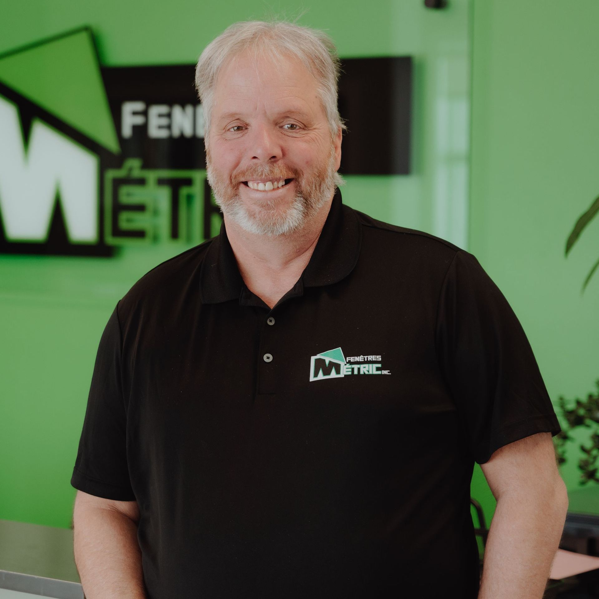 Homme aux cheveux gris et à la barbe en chemise noire avec un logo, souriant devant un mur vert avec une enseigne commerciale.