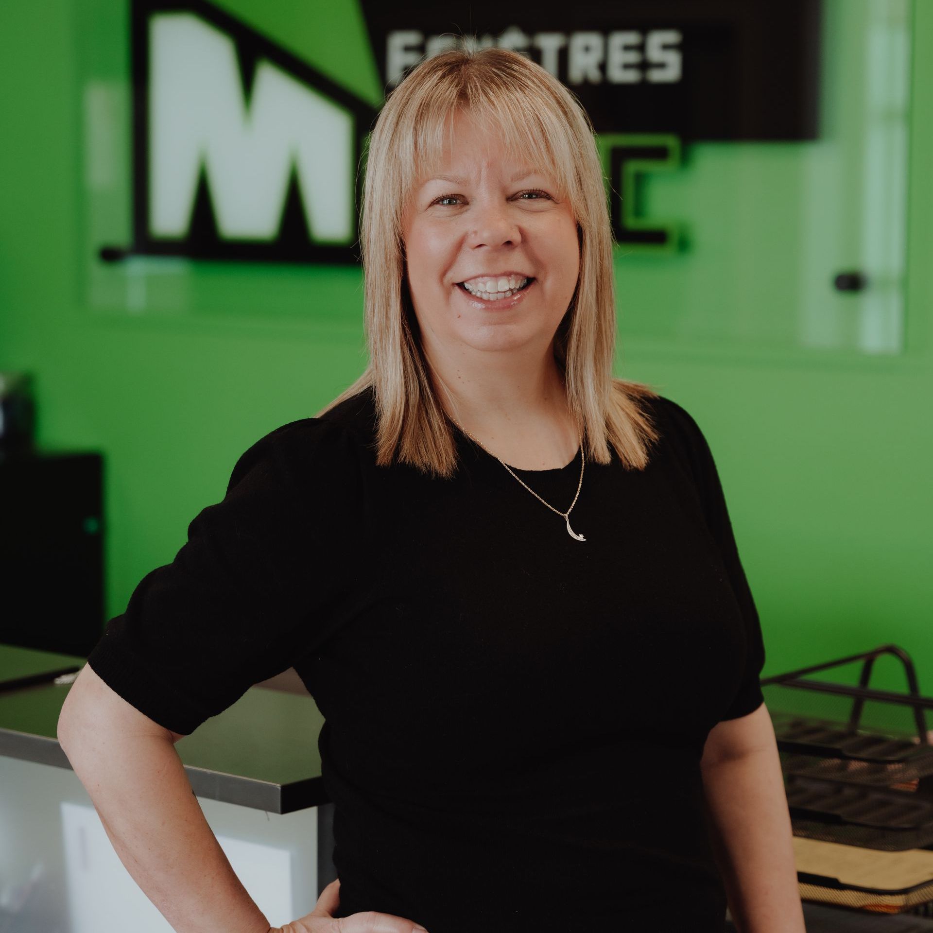 Une femme en haut noir sourit, appuyée contre un comptoir, dans un bureau au thème vert.