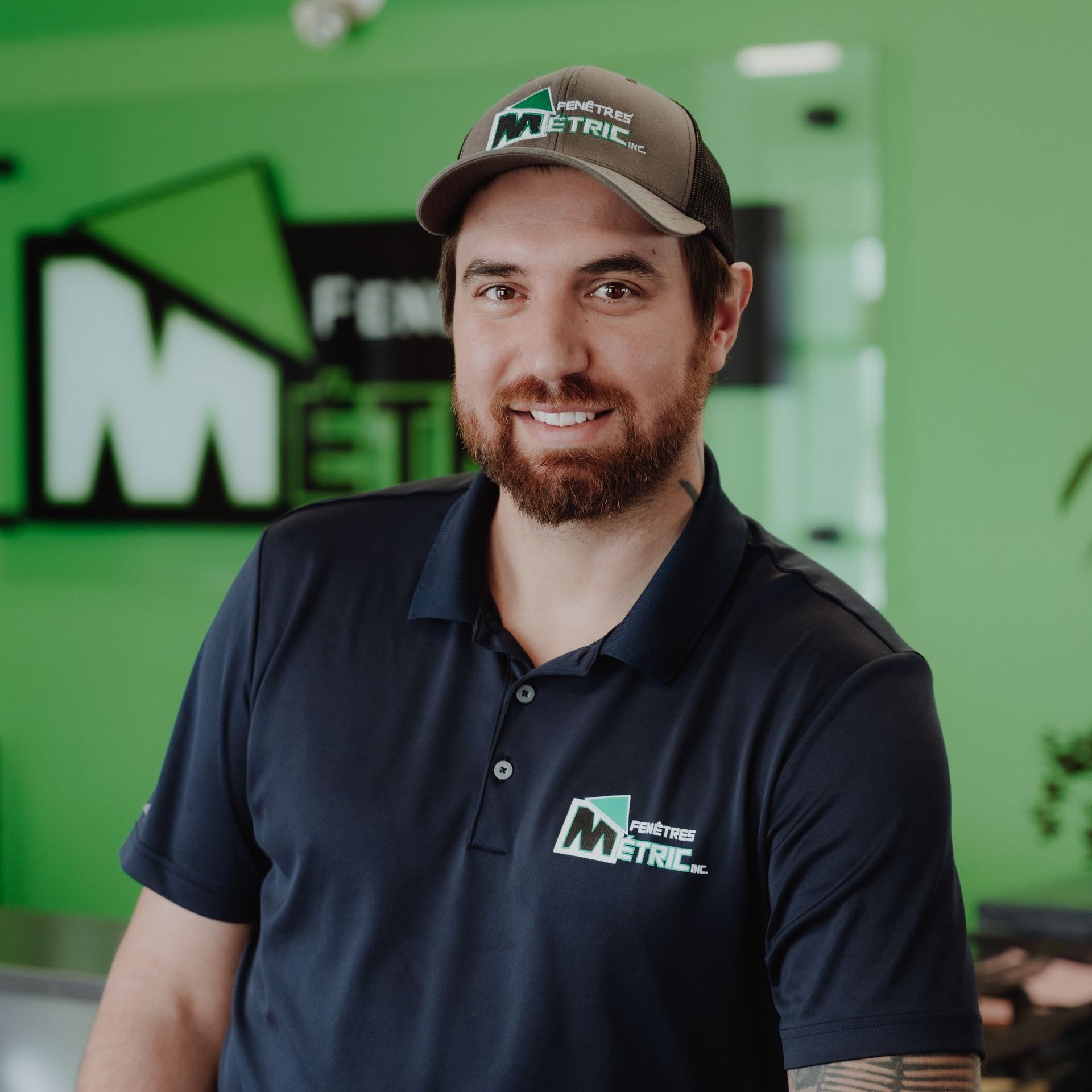 Un homme portant une casquette et un polo, souriant dans un bureau avec un logo d'entreprise.