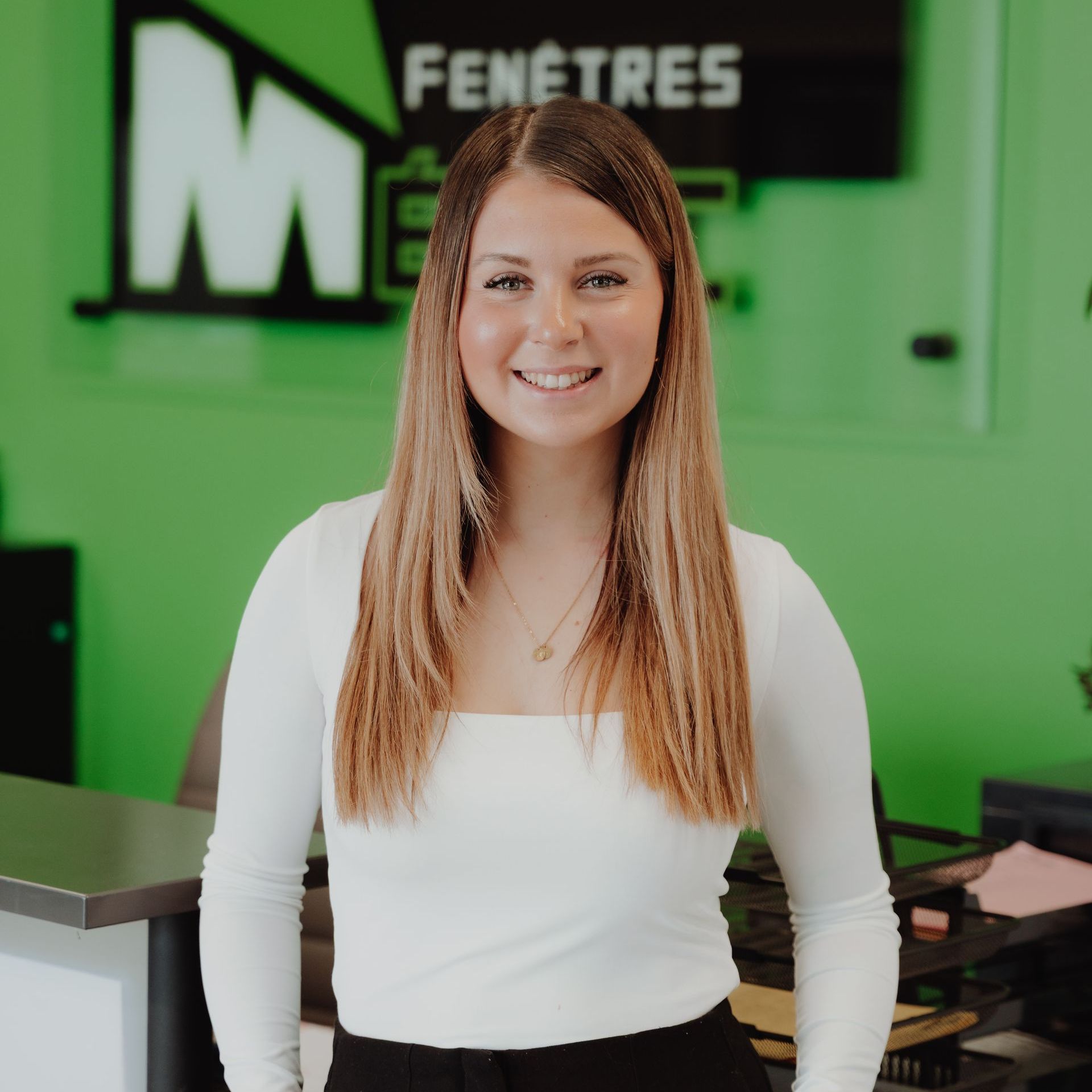 Une femme aux longs cheveux bruns sourit à la caméra devant un panneau vert sur lequel est écrit « Fenêtres Métric ».