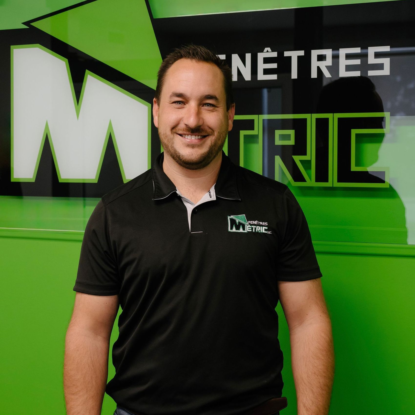 Un homme en polo noir sourit devant un logo Metric Windows vert.